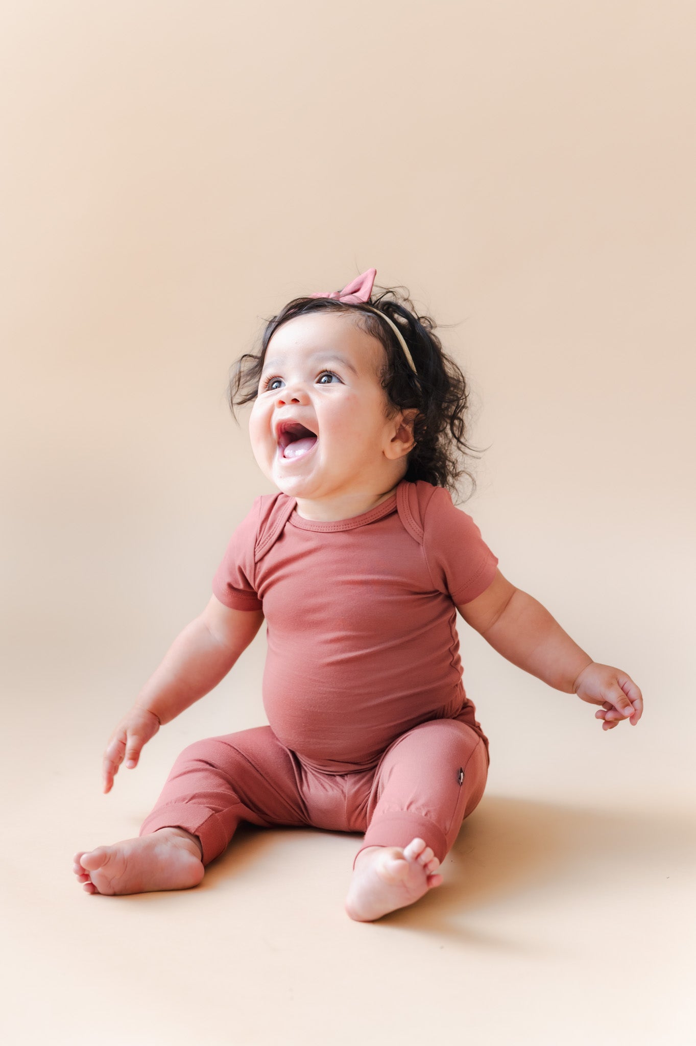 Baby wearing Pant in Ember over similar color bodysuit and bow headband on a beige background