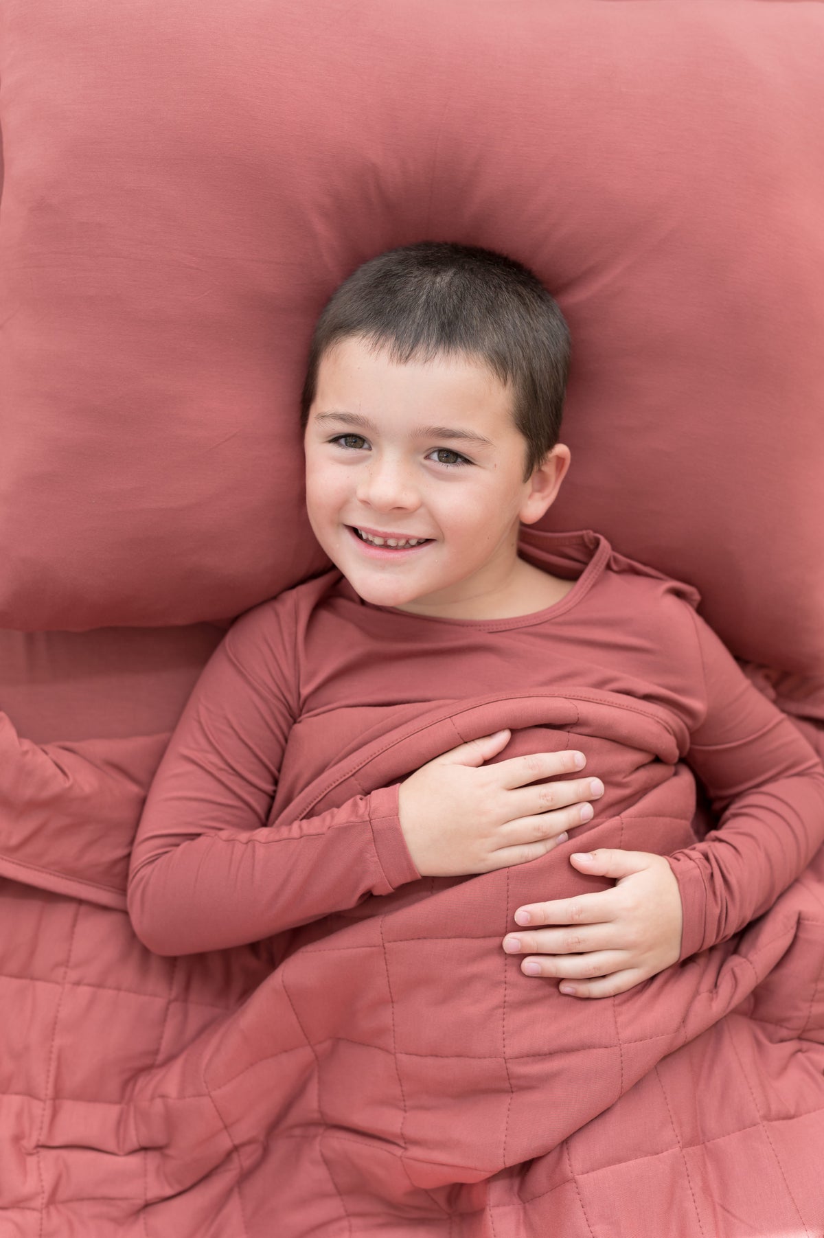 Boy lying on Standard Pillowcase in Ember covered under similar color blanket while wearing a similar color shirt