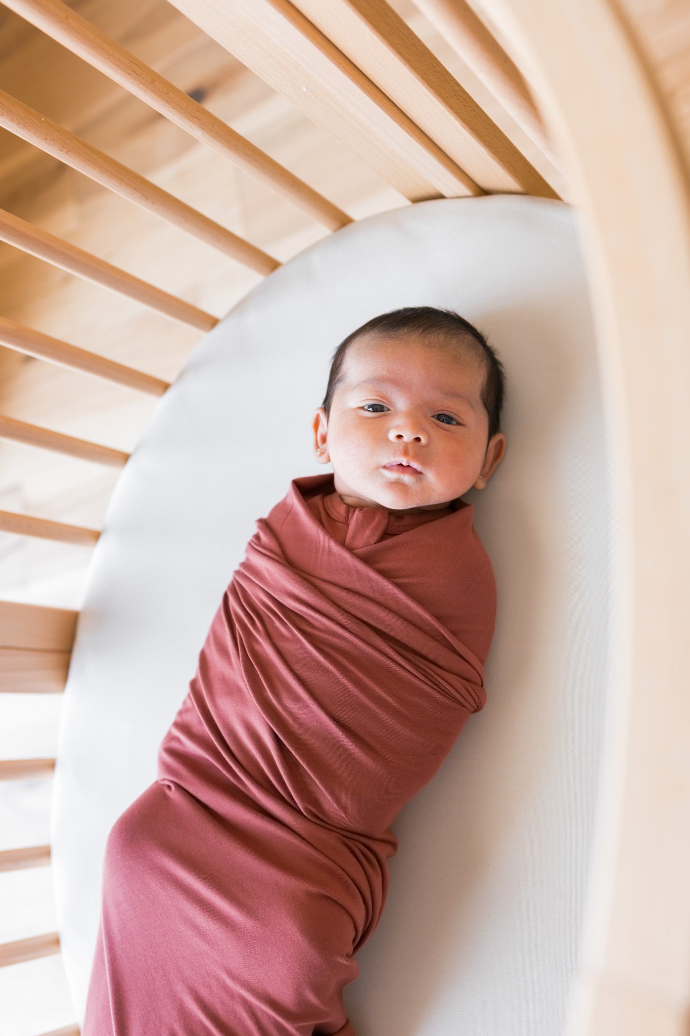 A newborn baby swaddled in a dusty terracotta wrap blanket, lying on a white mattress inside a light wood slatted crib.