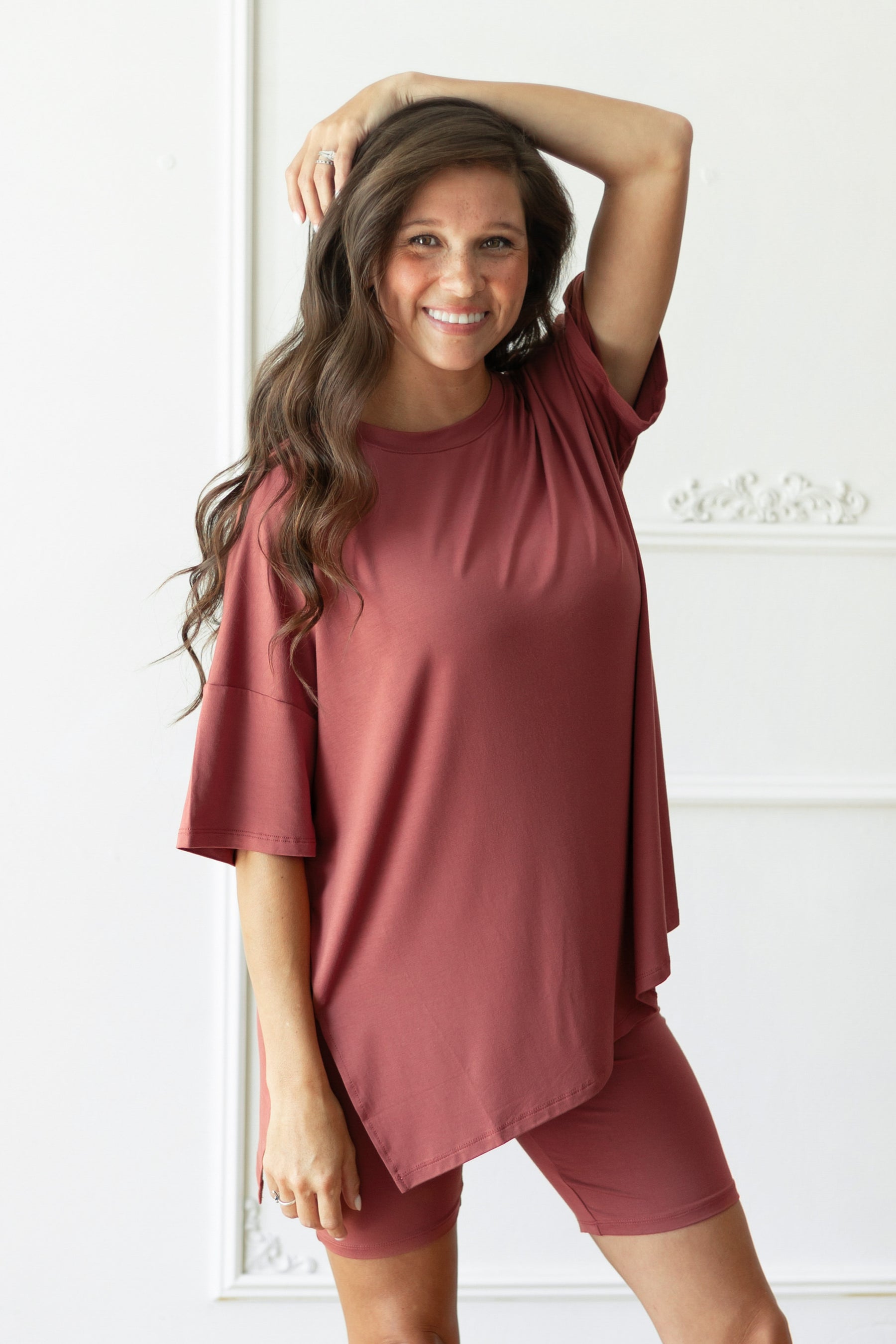 A woman modeling a matching dusty terracotta short-sleeve t-shirt and fitted biker shorts set against a white paneled wall.
