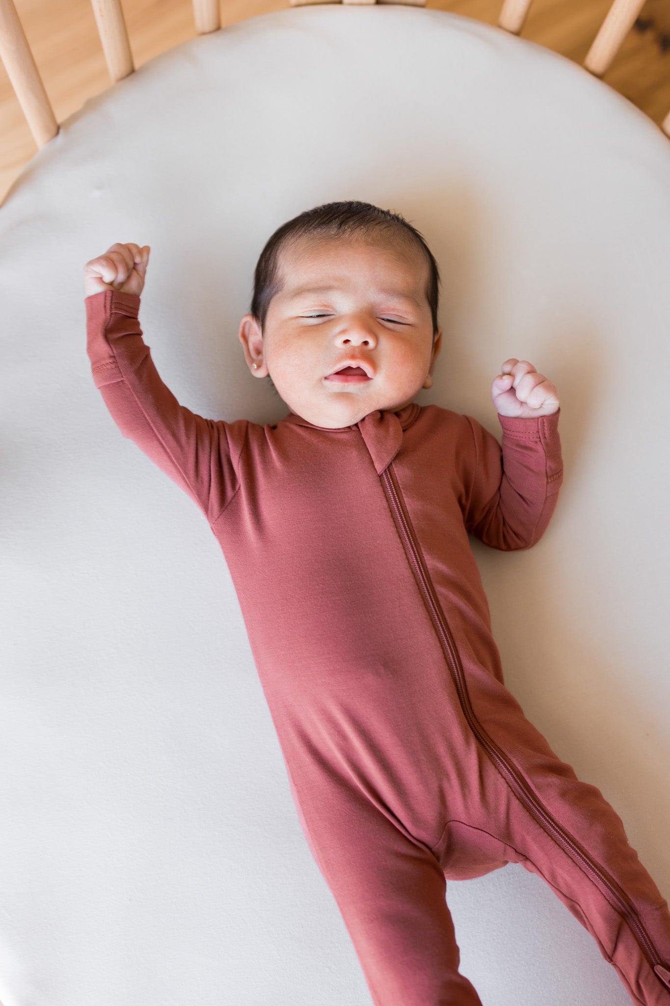 A newborn baby wearing a dusty terracotta zip-up sleeper, lying on their back inside a light-colored wooden crib.