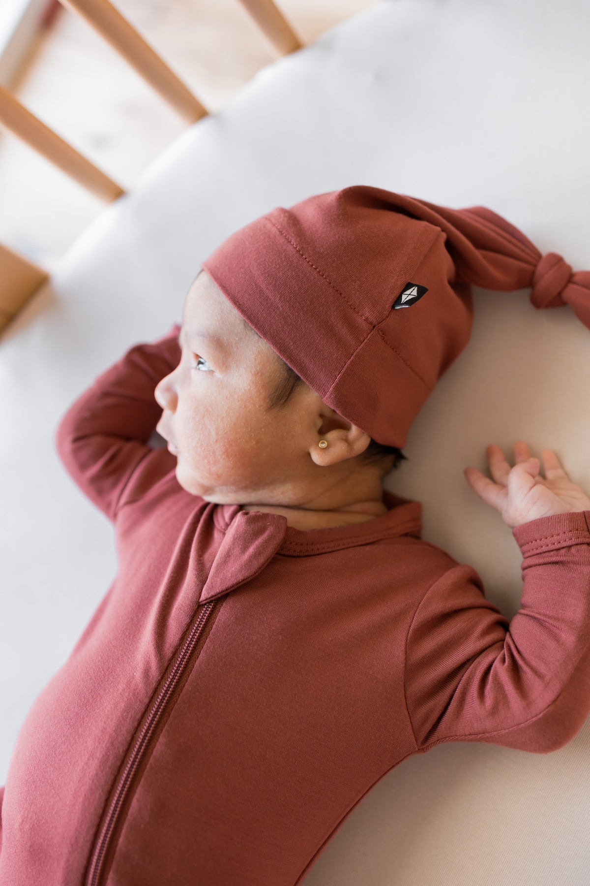 A newborn baby wearing a dusty rose terracotta long-sleeve zip-up sleeper and matching knotted beanie hat, lying on a white mattress in a crib.