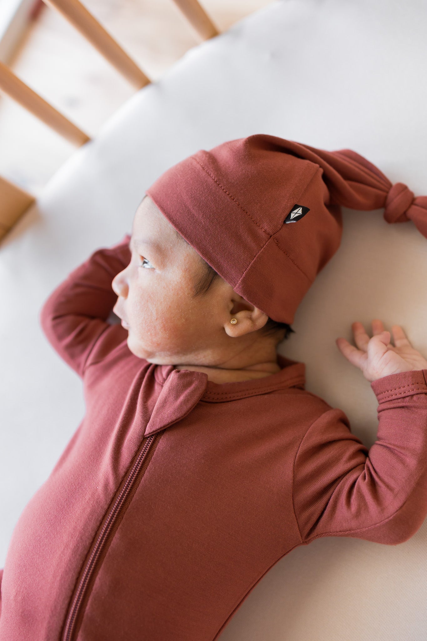 A newborn baby wearing a dusty rose terracotta long-sleeve zip-up sleeper and matching knotted beanie hat, lying on a white mattress in a crib.