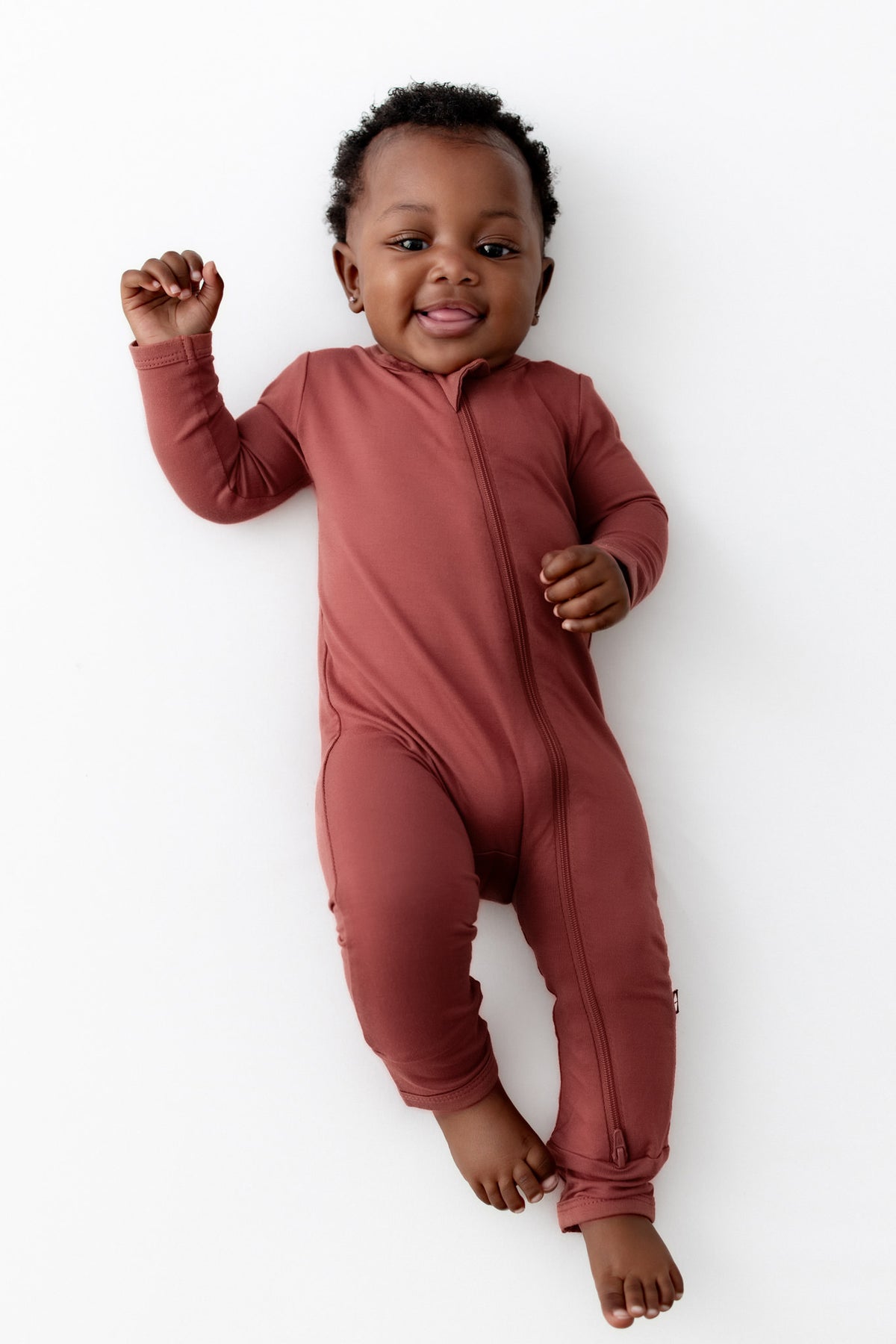 A baby lying on their back wearing a long-sleeve, one-piece zip-up romper in a muted terracotta color, set against a plain white background.