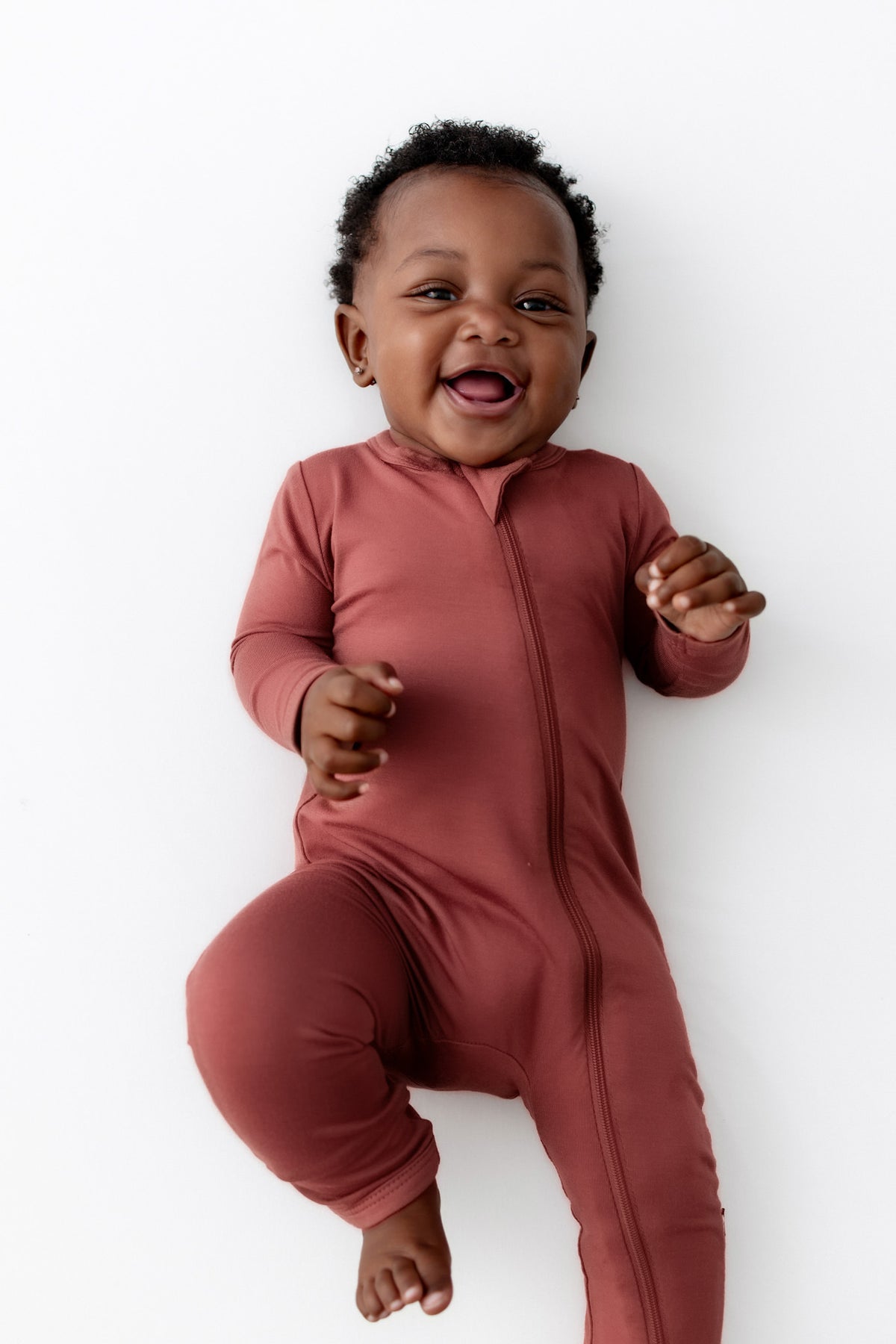 A smiling baby wearing a terracotta zip-up footless romper, lying on a clean white background.