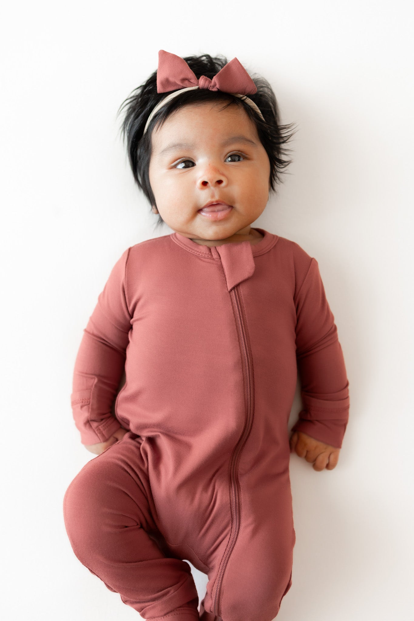 A baby wearing a dusty terracotta long-sleeve footless romper with a front zipper and a matching bow headband, lying on a white background.
