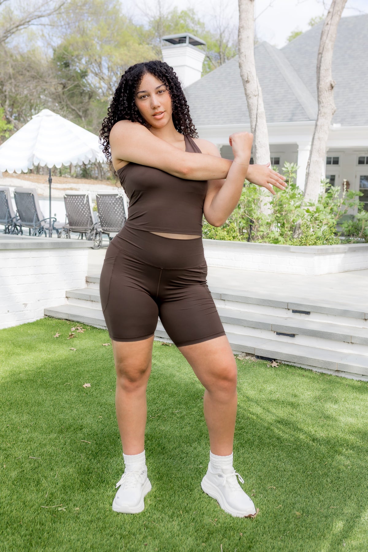Female model standing in the grass in front of a house stretching wearing the Bamboo Flex High Rise Biker Short in Espresso and matching bra tank