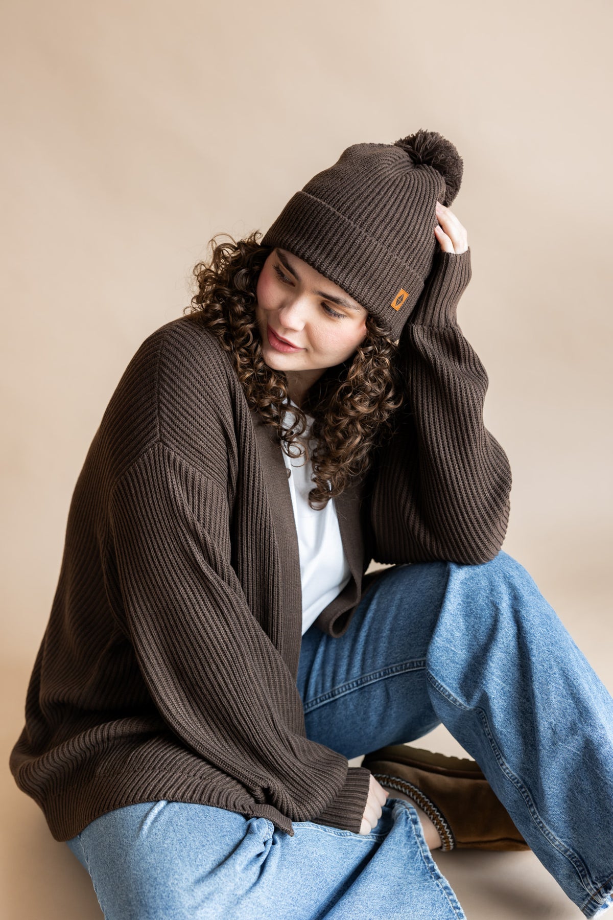 Modal posing in Chunky Knit Adult Pom in Espresso over Chunky knit Cardigan and blue jeans on a beige background
