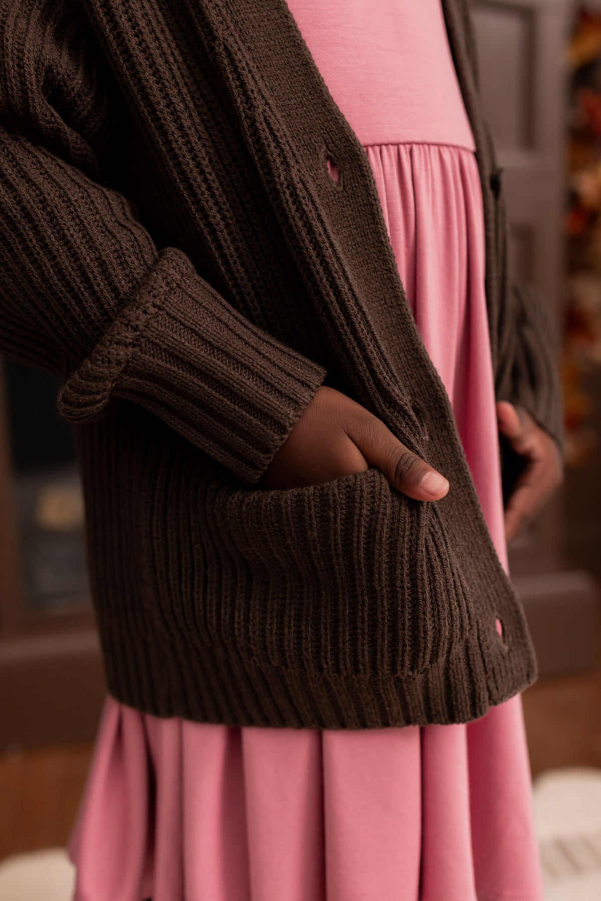 Close up of Girl wearing Chunky Knit Oversized Cardigan in Espresso over Twirl dress in pink standing in a living room