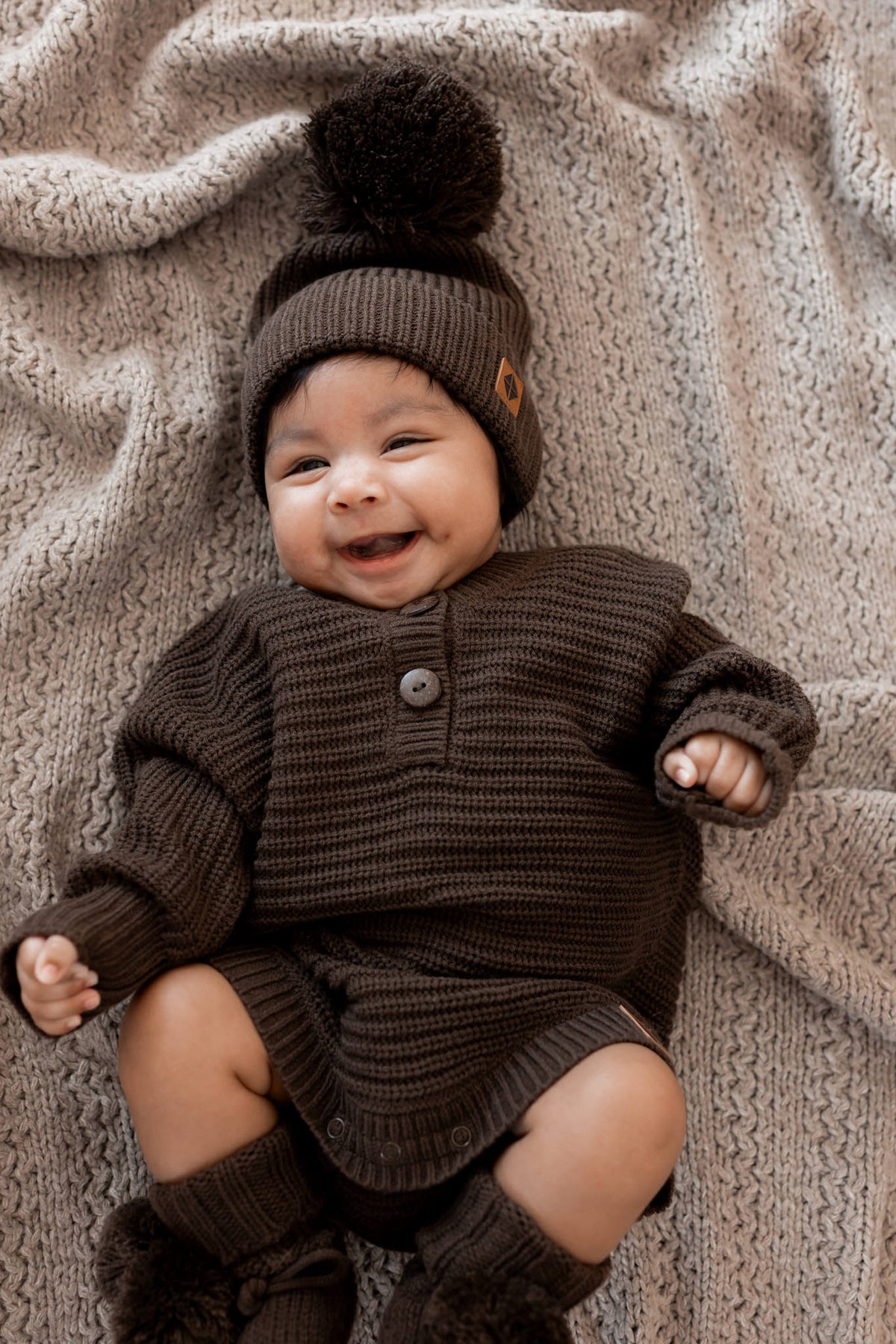 Baby lying on a beige blanket wearing Chunky Knit Pom Beanie, chunky knit romper and knit booties in Espresso
