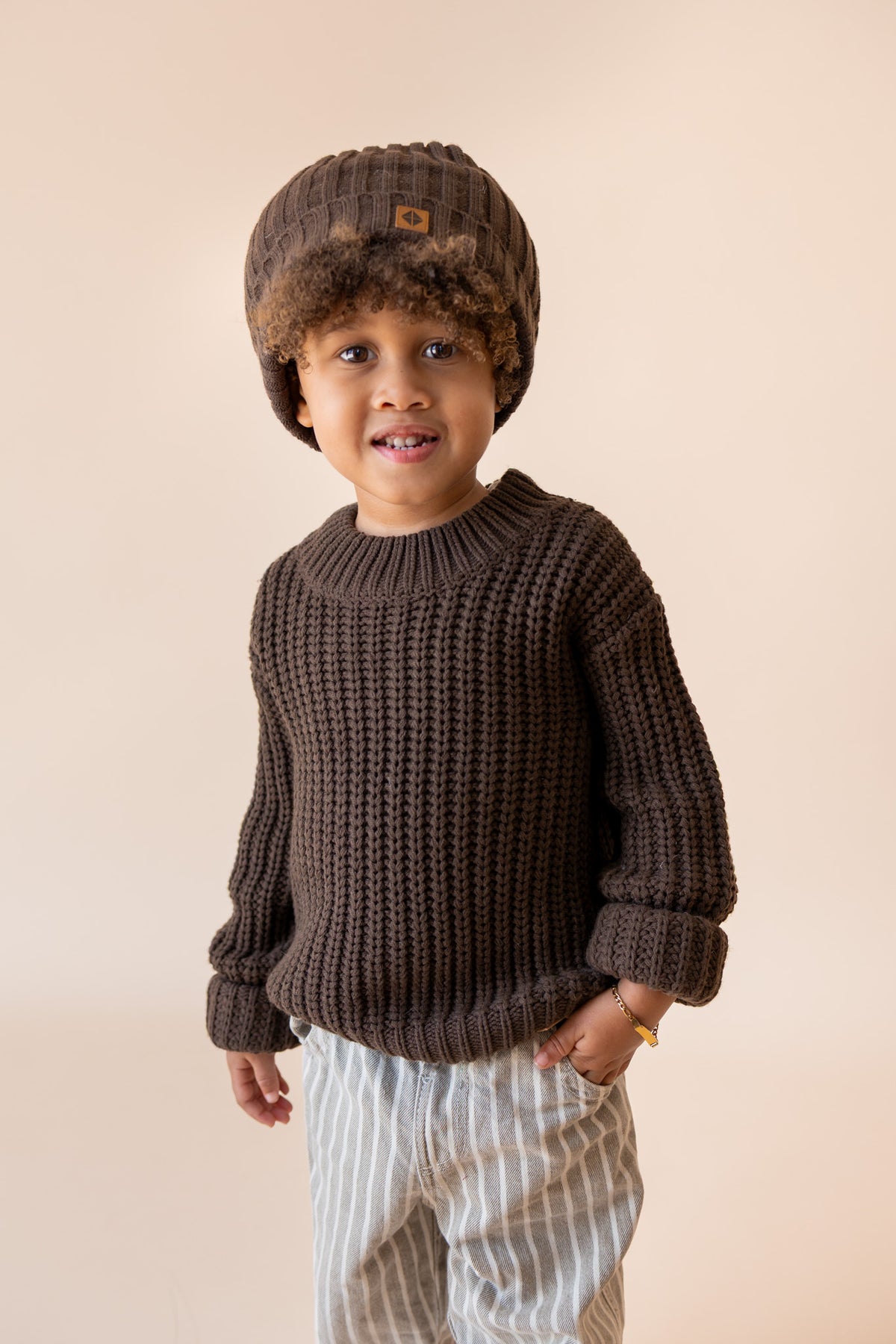 Boy wearing Chunky Knit Sweater and chunky knit ribbed beanie in espresso over pants on a beige background