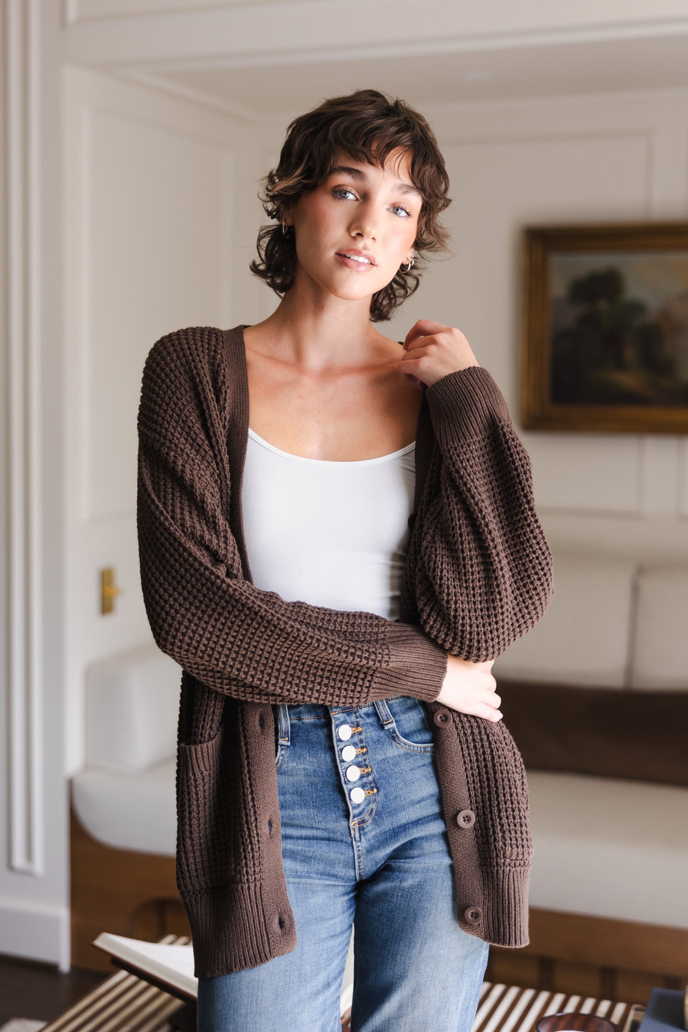 Modal standing in a living room while wearing Chunky Knit Women's Waffle Cardigan in Espresso over white camisole and blue jeans