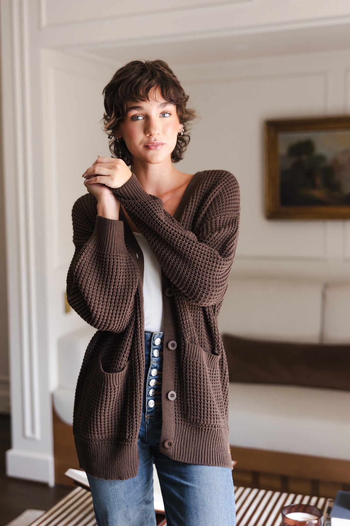Modal posing in Chunky Knit Women's Waffle Cardigan in Espresso over white camisole and blue jeans while standing in a living room