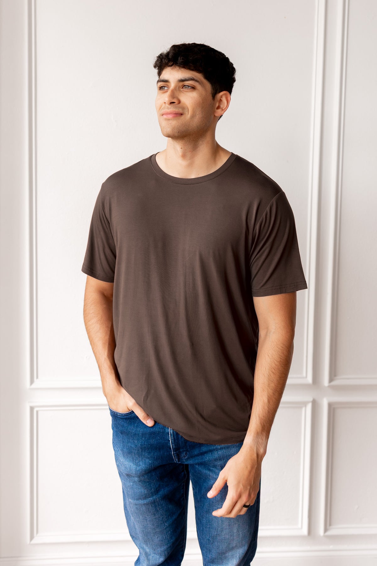 Man wearing a brown t-shirt and blue jeans standing against a white paneled wall.