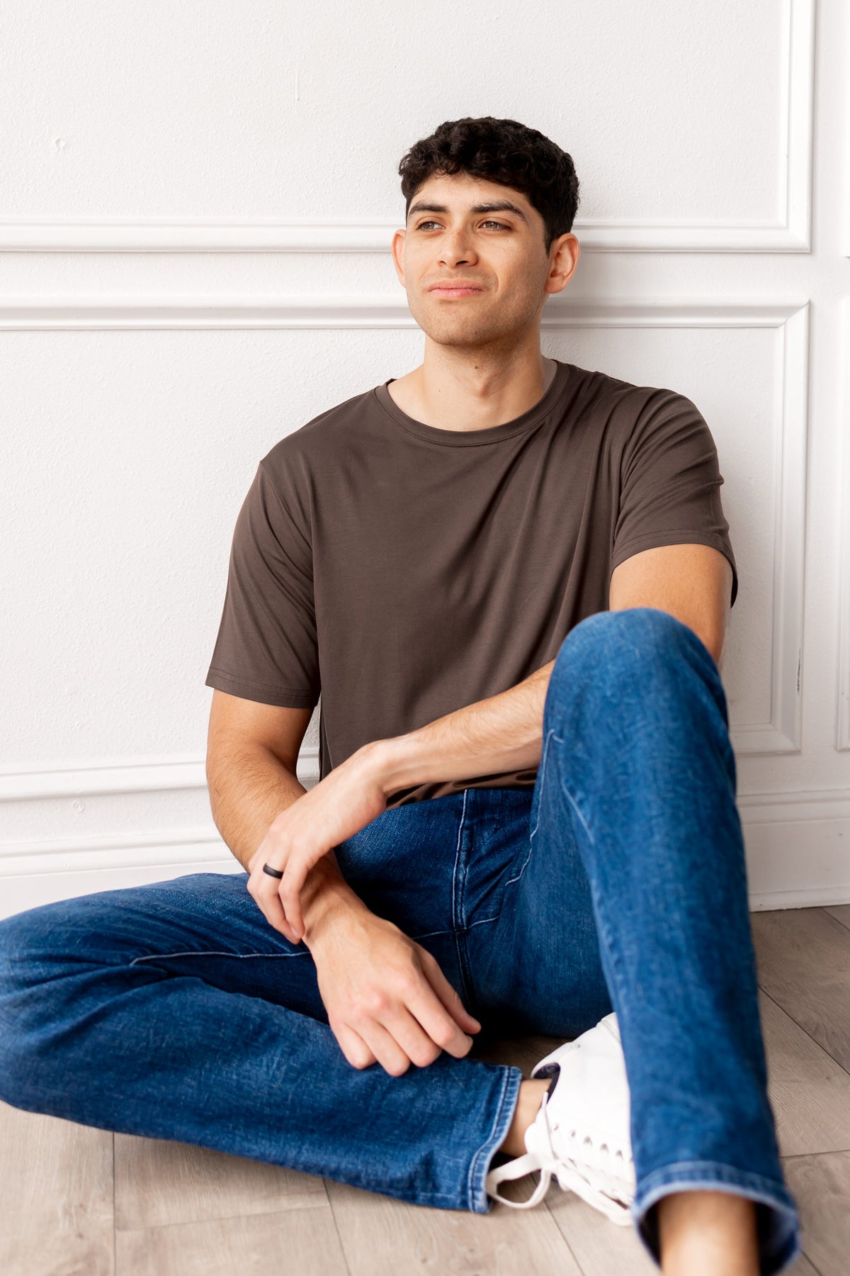 Man sitting on a floor wearing a brown t-shirt and blue jeans.