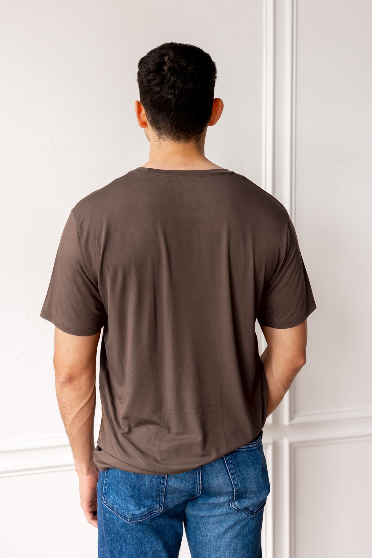 Man wearing a brown t-shirt and blue jeans standing against a white wall.