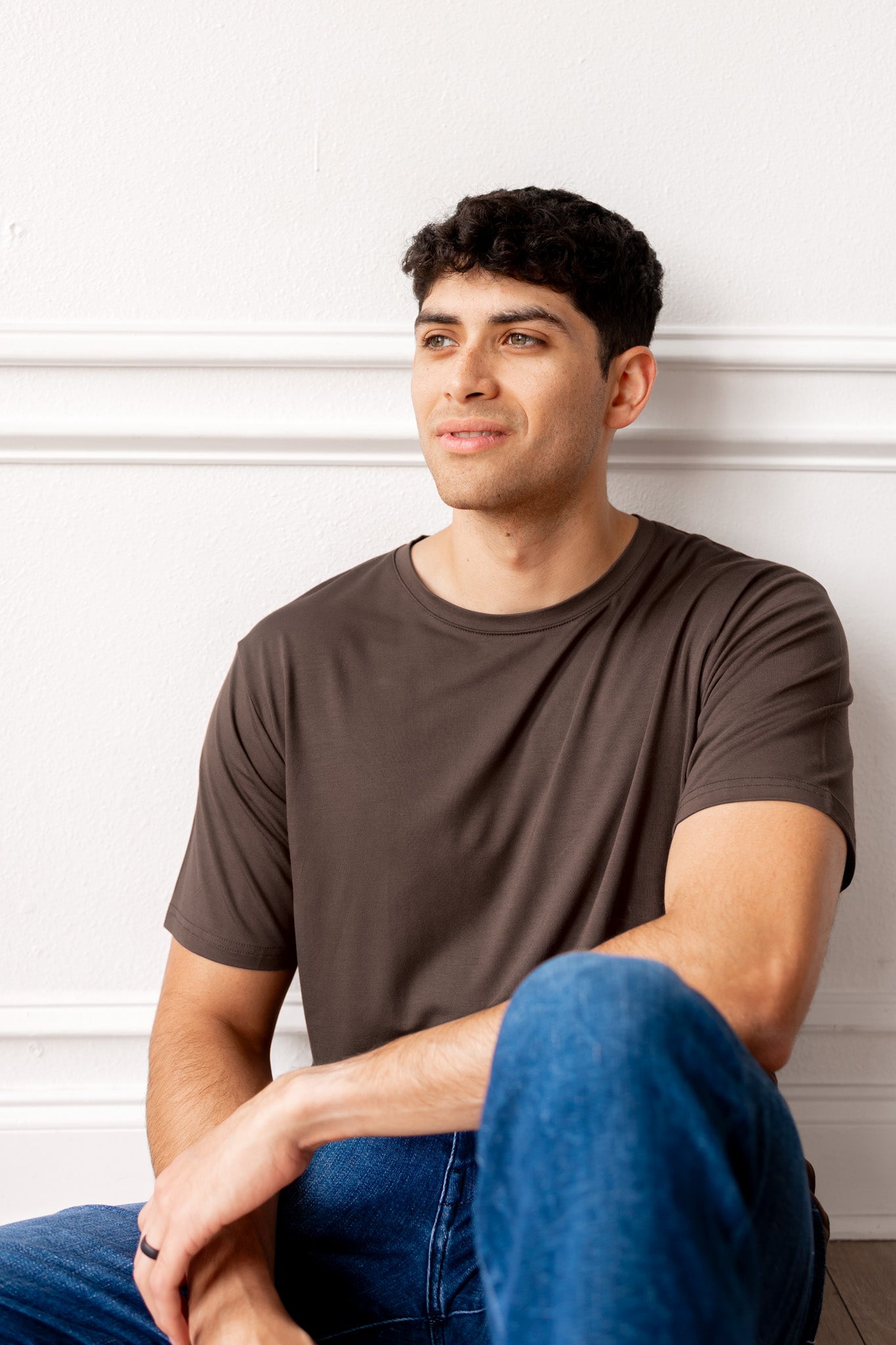 Man sitting on a floor wearing a brown t-shirt and blue jeans a white paneled wall.