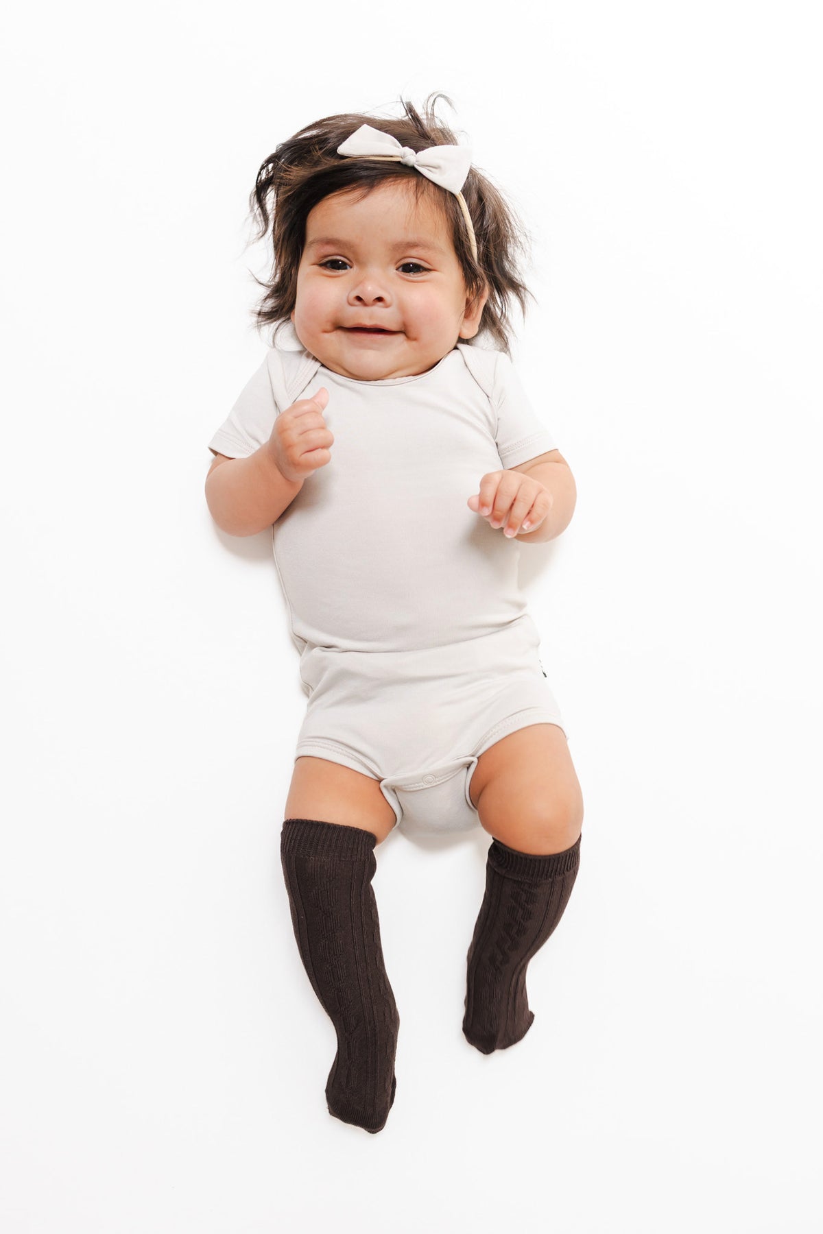A smiling baby lying on a white background, wearing a light gray short-sleeve bodysuit, dark brown knee-high socks, and a white bow headband.