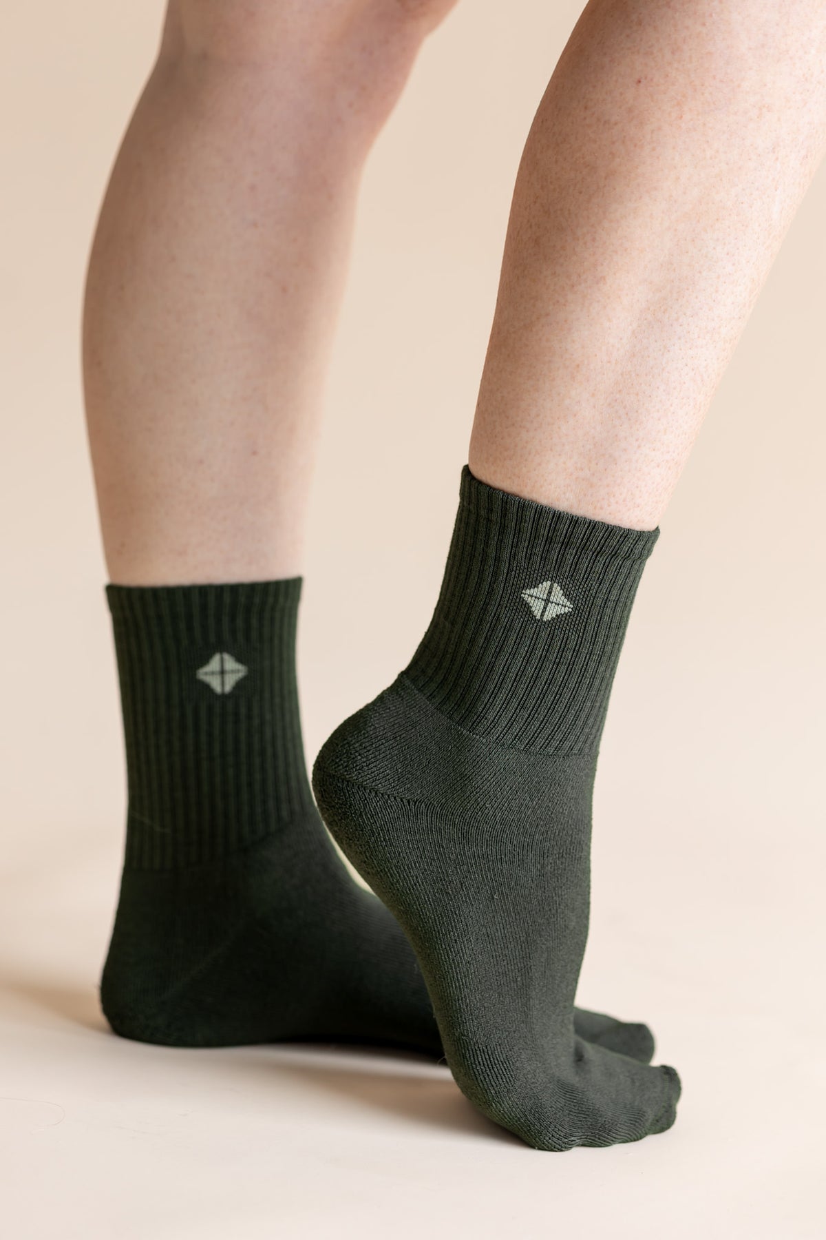 A pair of dark forest green crew socks worn on a person's feet, shown against a solid light beige background. The socks feature a ribbed cuff and a small white geometric logo on the front.
