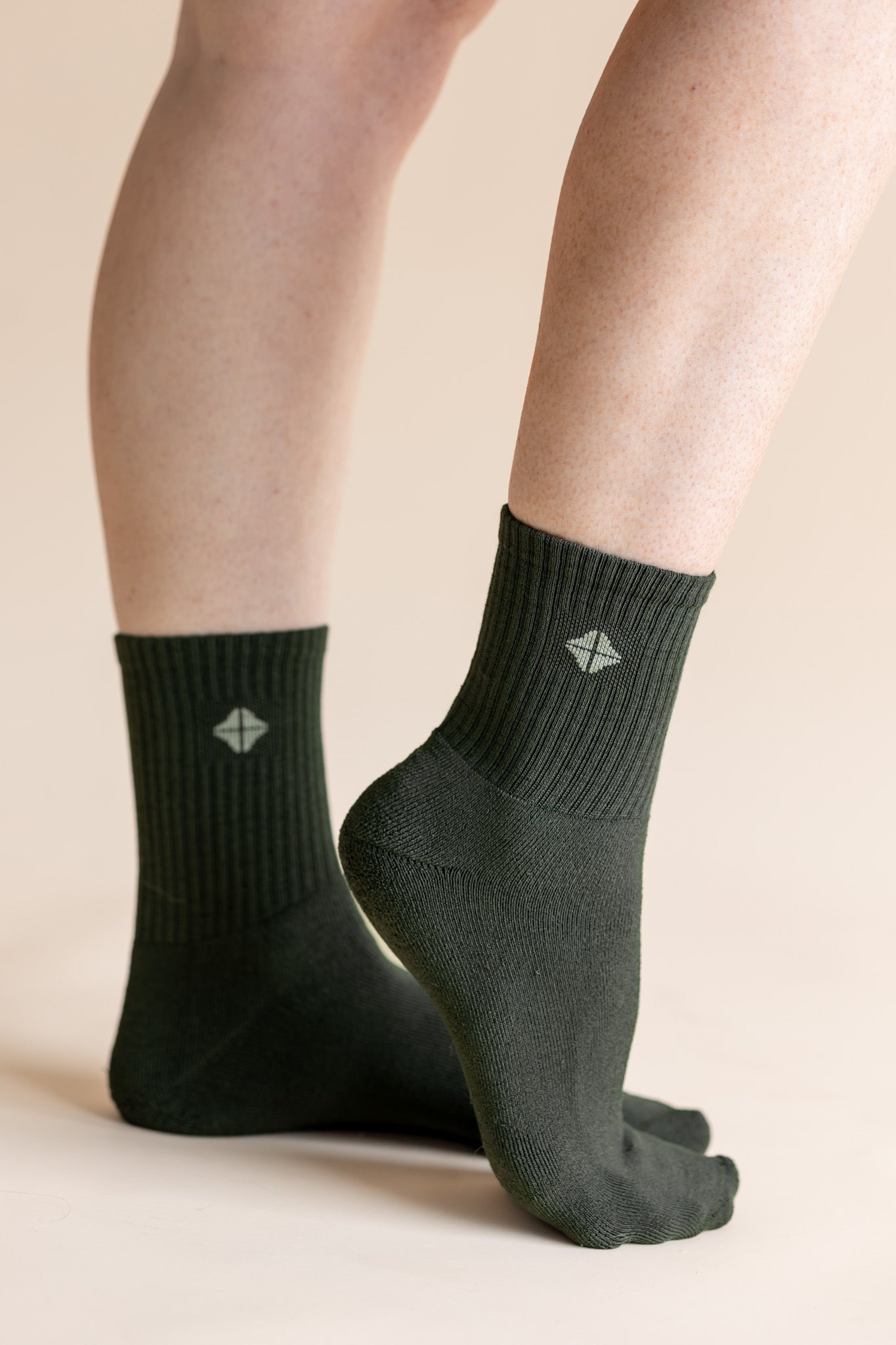 A pair of dark forest green crew socks worn on a person's feet, shown against a solid light beige background. The socks feature a ribbed cuff and a small white geometric logo on the front.