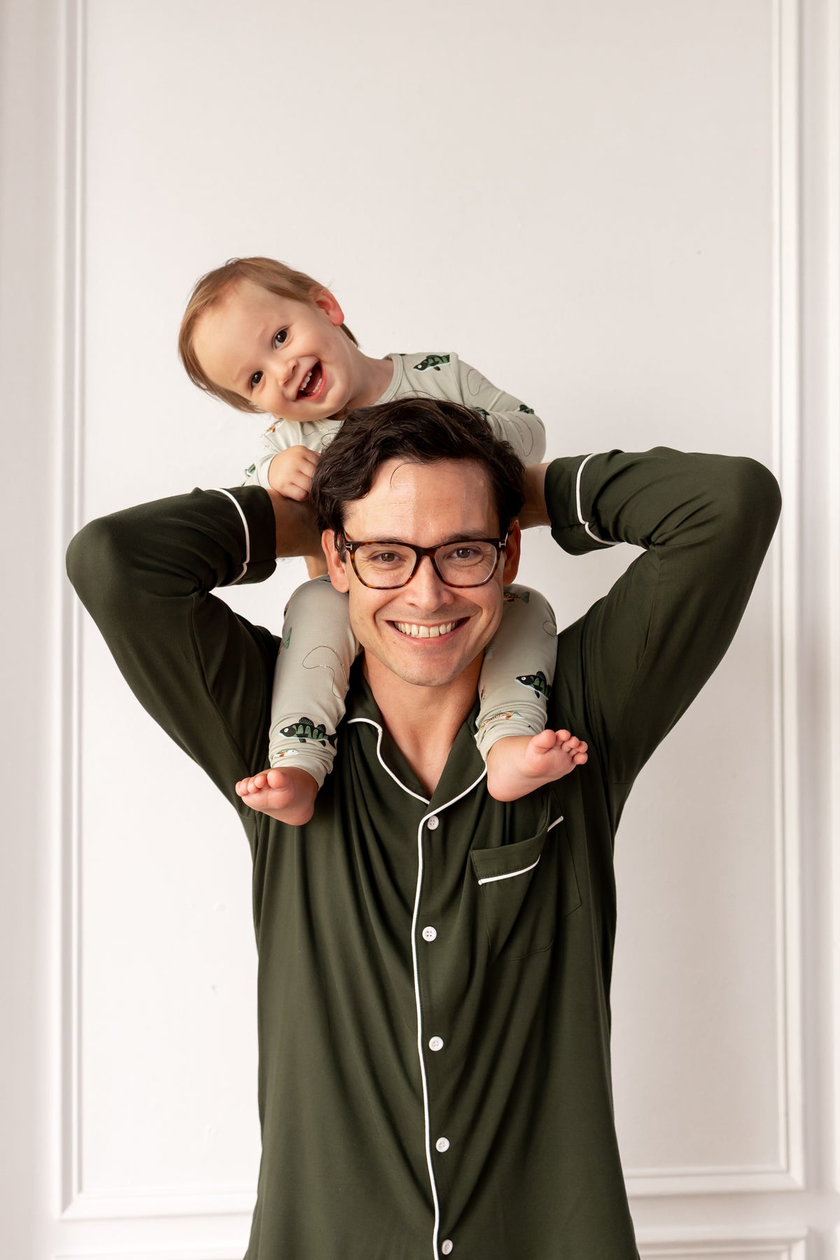 Man in green pajamas holding a baby on his shoulders against a white wall.