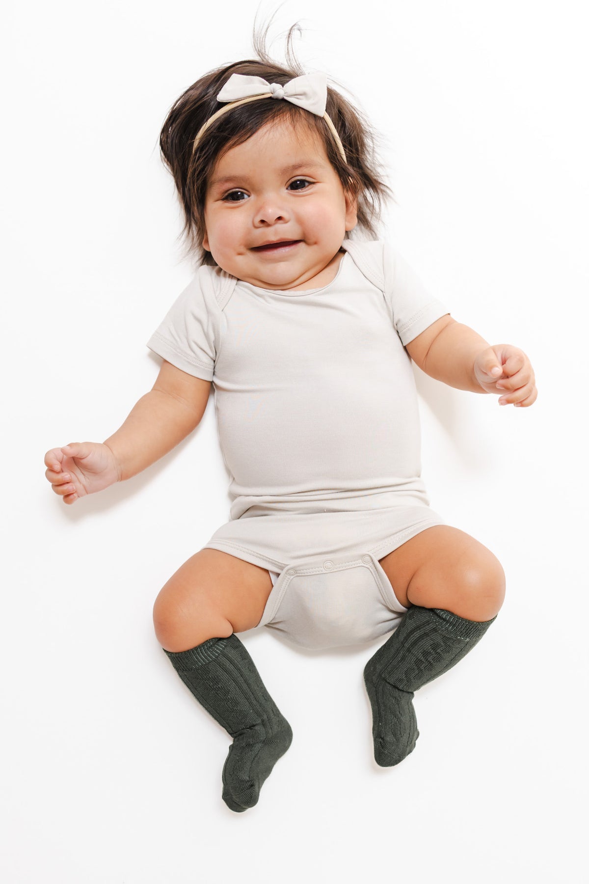 A baby lying on a white background wearing a light beige short-sleeve romper, dark green knee-high socks, and a matching beige bow headband.