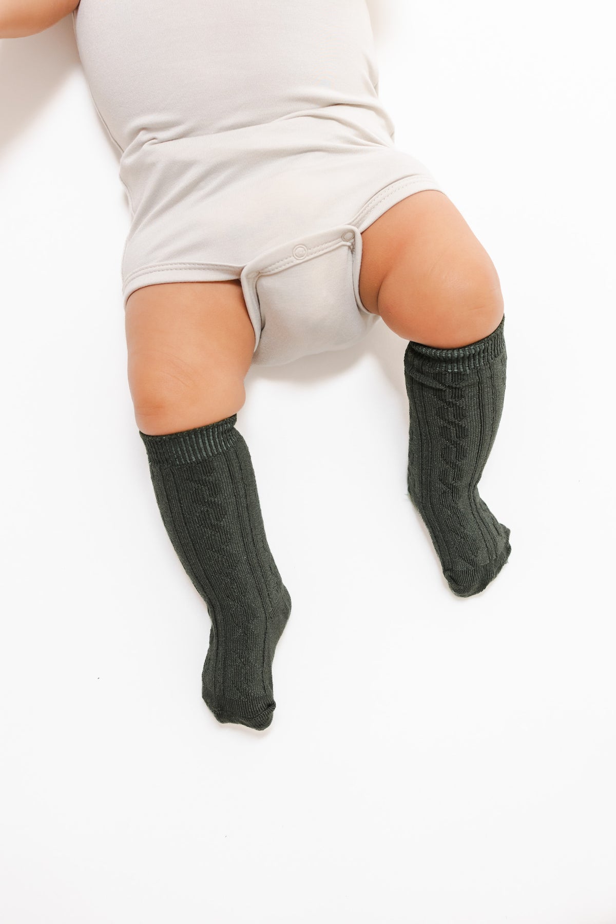 A baby lying on a white surface wearing dark green cable-knit knee-high socks and a light beige bodysuit.