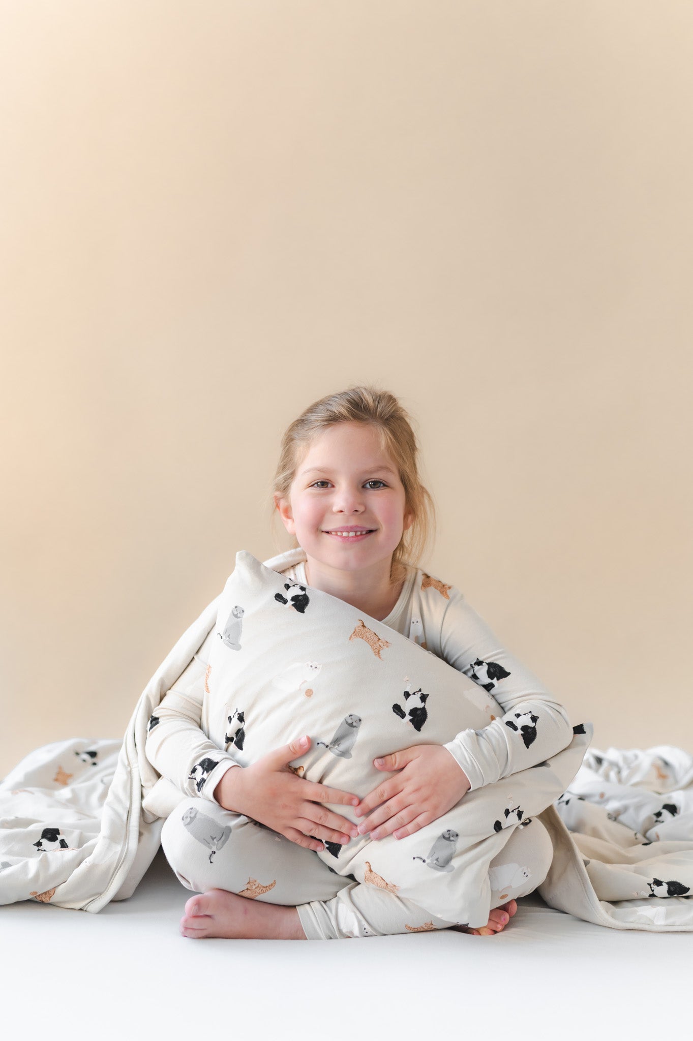 Child wrapped in a soft blanket with cat patterns and holding a matching pillow on a plain background