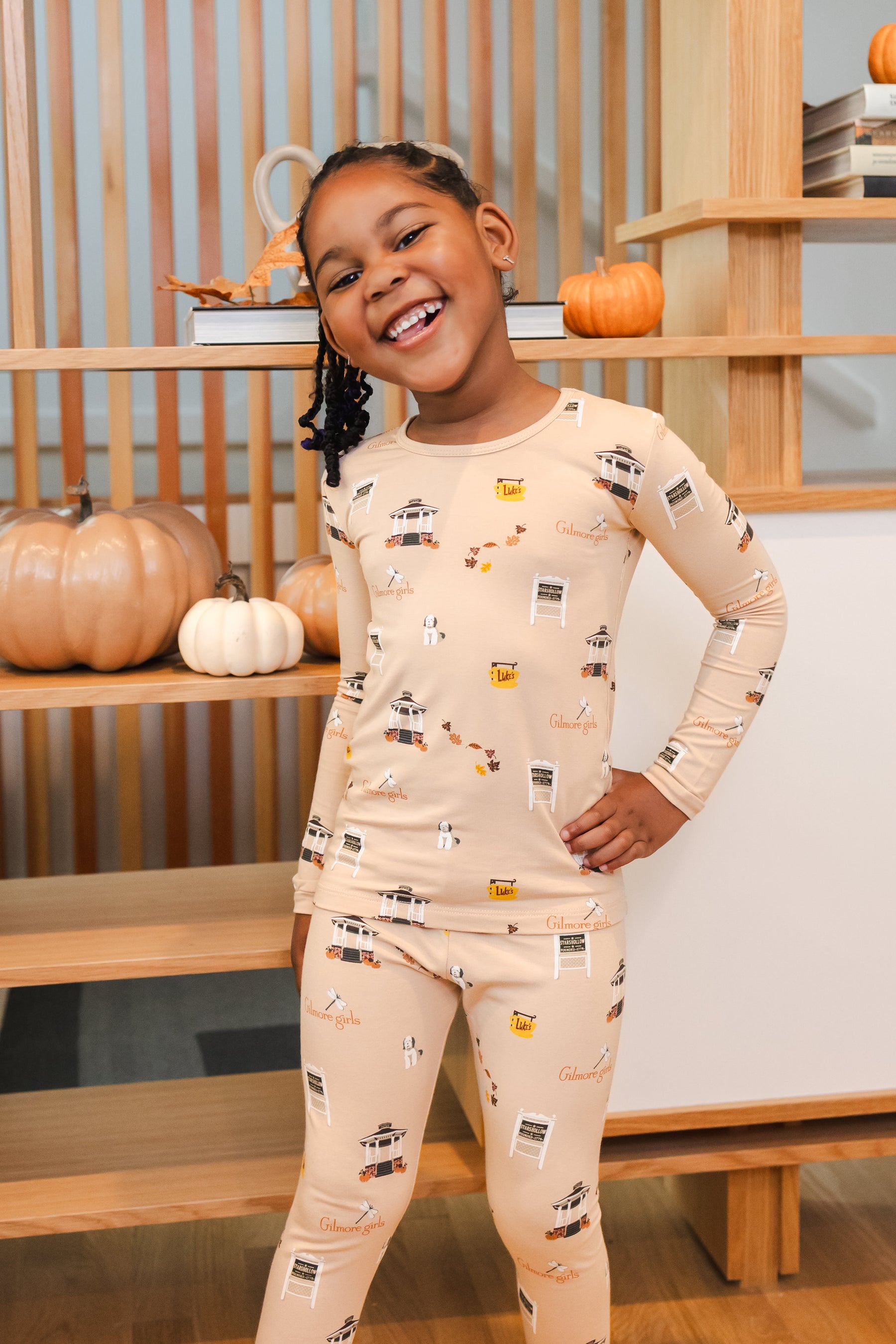 Child wearing pajamas with a Gilmore Girls pattern in a room with wooden shelves and pumpkins.