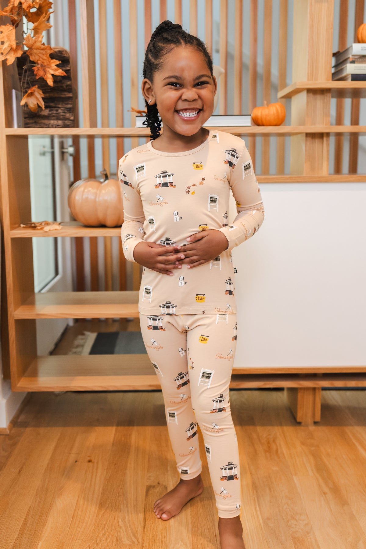 Child wearing pajamas with Gilmore Girls pattern standing in a room with wooden shelves and pumpkins.