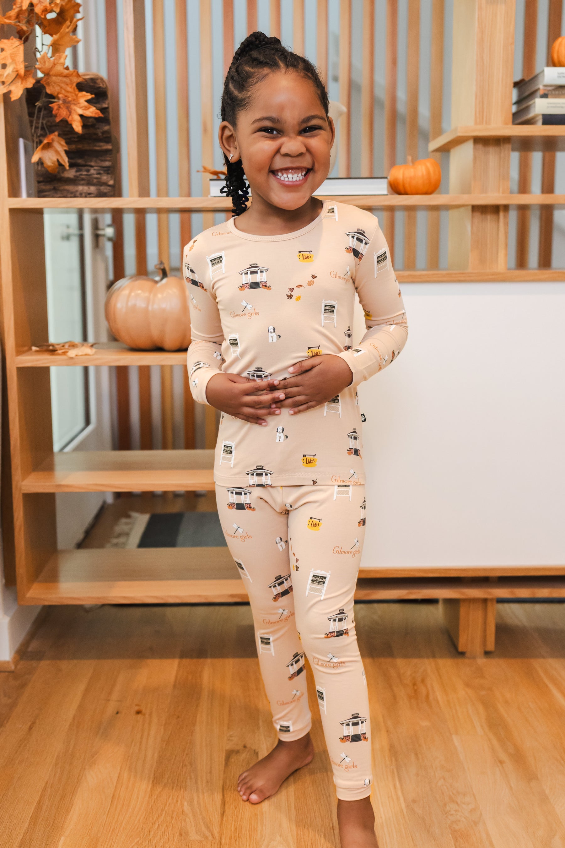 Child wearing pajamas with Gilmore Girls pattern standing in a room with wooden shelves and pumpkins.