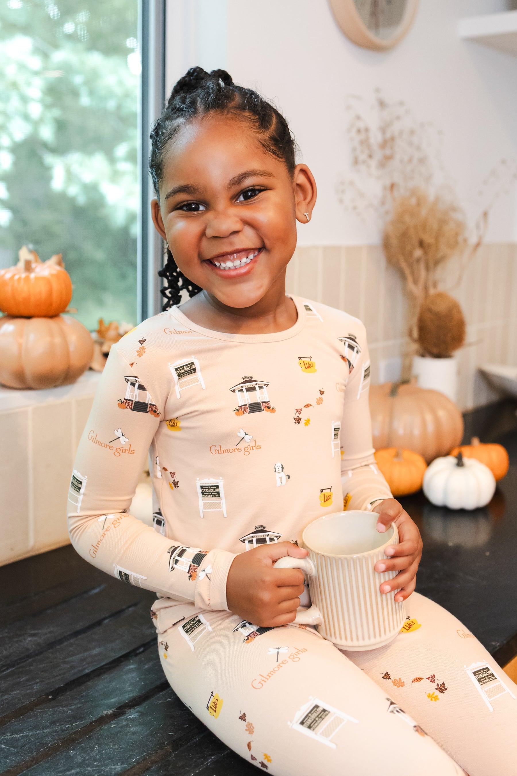 Child in pajamas with Gilmore Girls inspired pattern holding a mug, surrounded by pumpkins indoors.