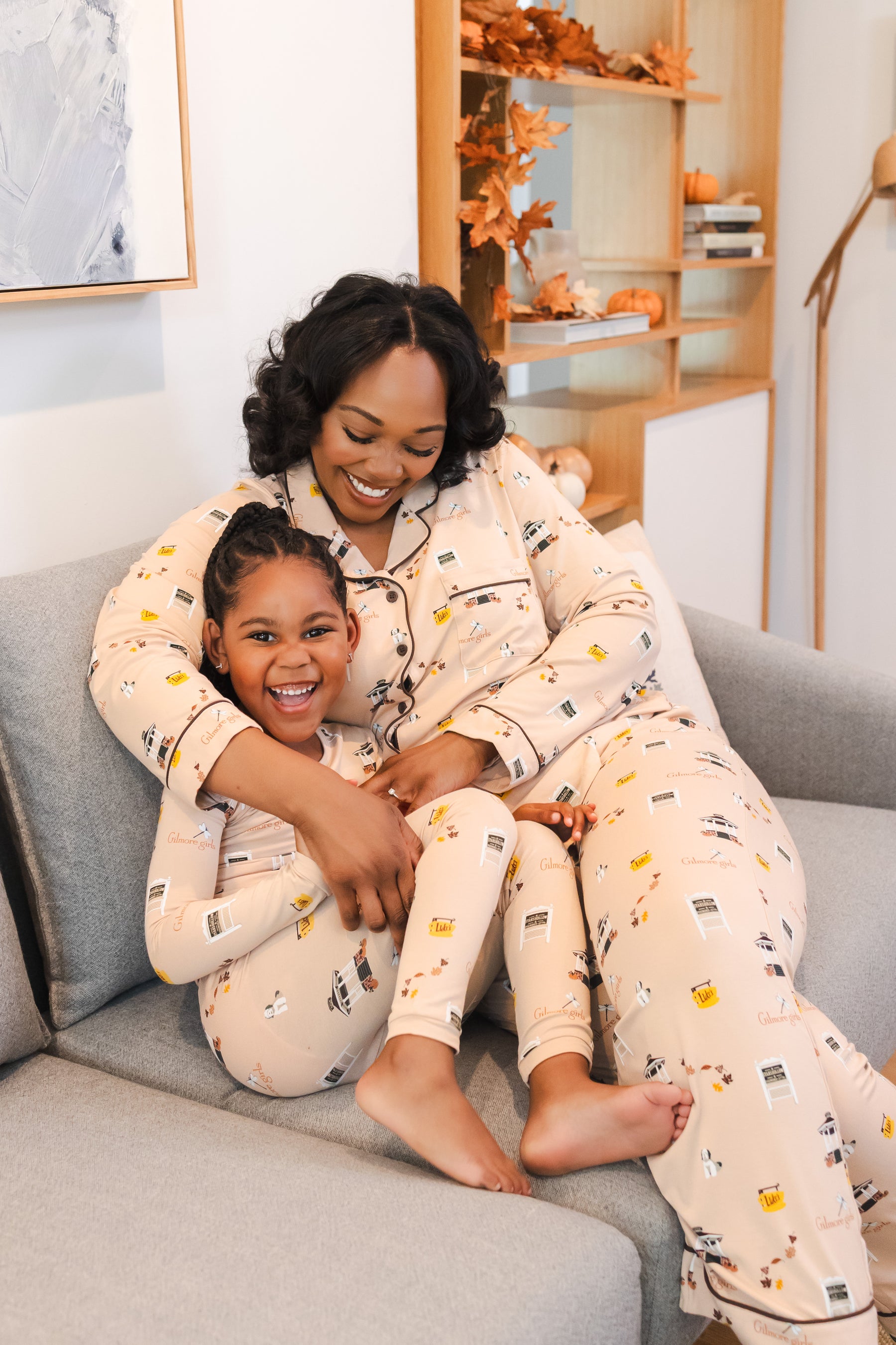 Woman and child sitting on a couch wearing matching Gilmore Girls pajamas with a fall-themed design.