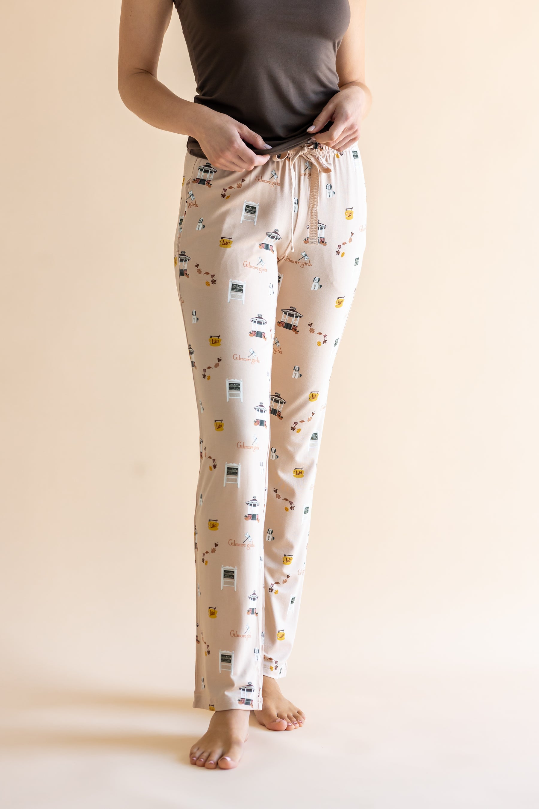 A person wearing cream-colored lounge pants with a Gilmore Girls inspired pattern, paired with a dark gray top, standing against a solid beige background.