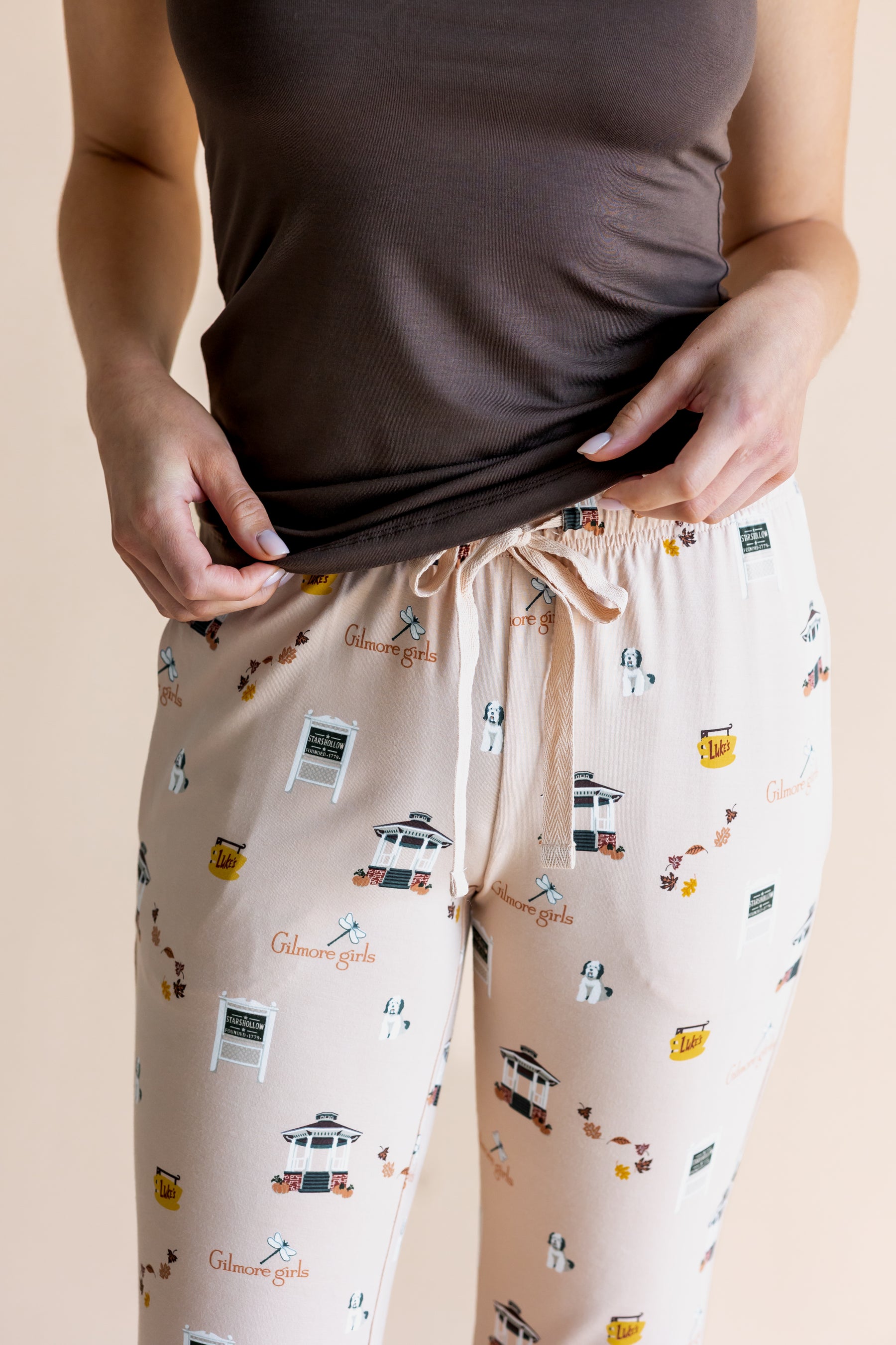 A person wearing beige drawstring pajama pants with a small multicolor Gilmore Girls themed print, paired with a dark brown sleeveless top, shown against a plain beige background.