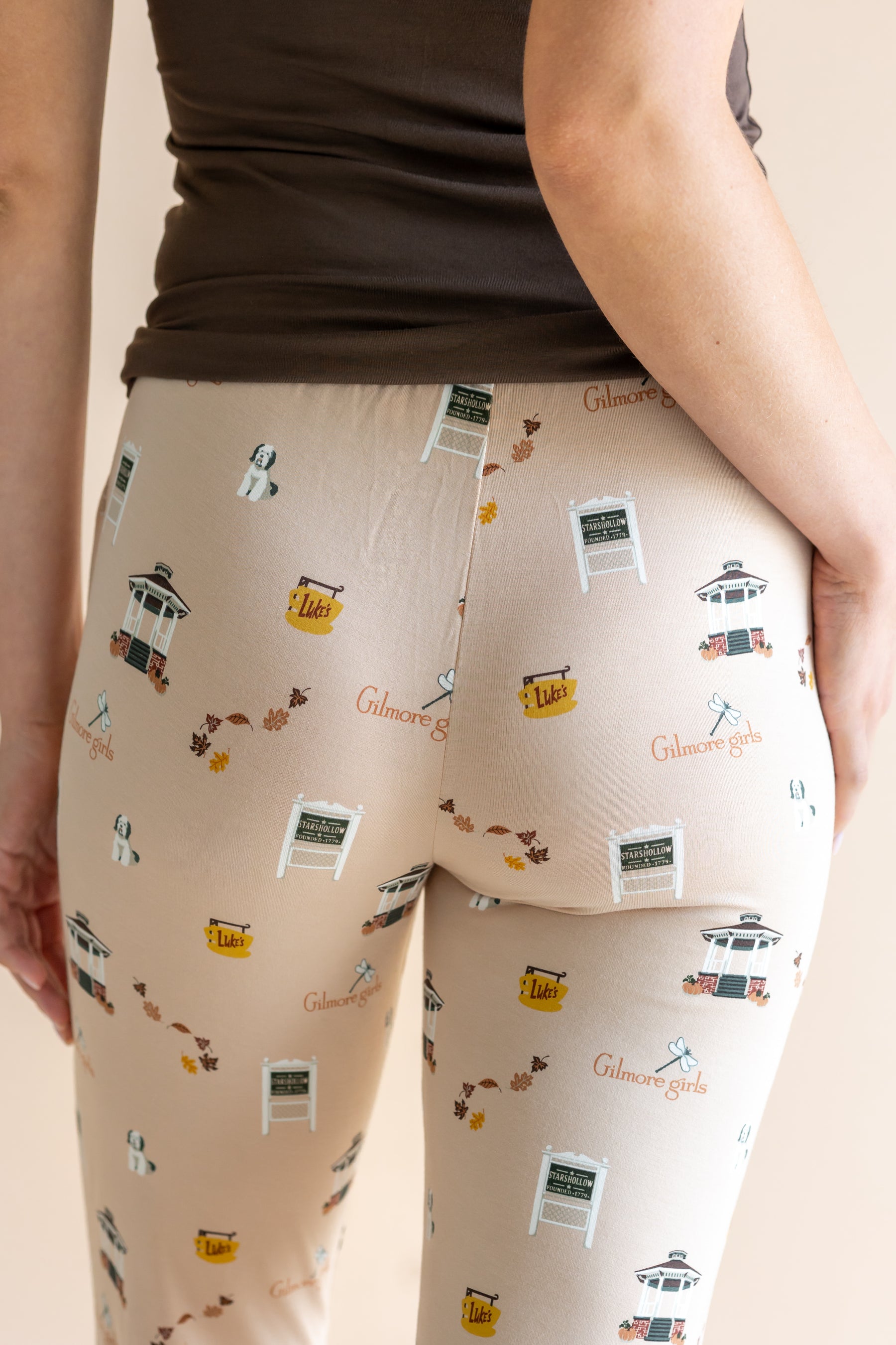 Close-up back view of a person wearing beige pants with a repeating print of Gilmore Girls elements, paired with a dark brown top against a plain beige background.