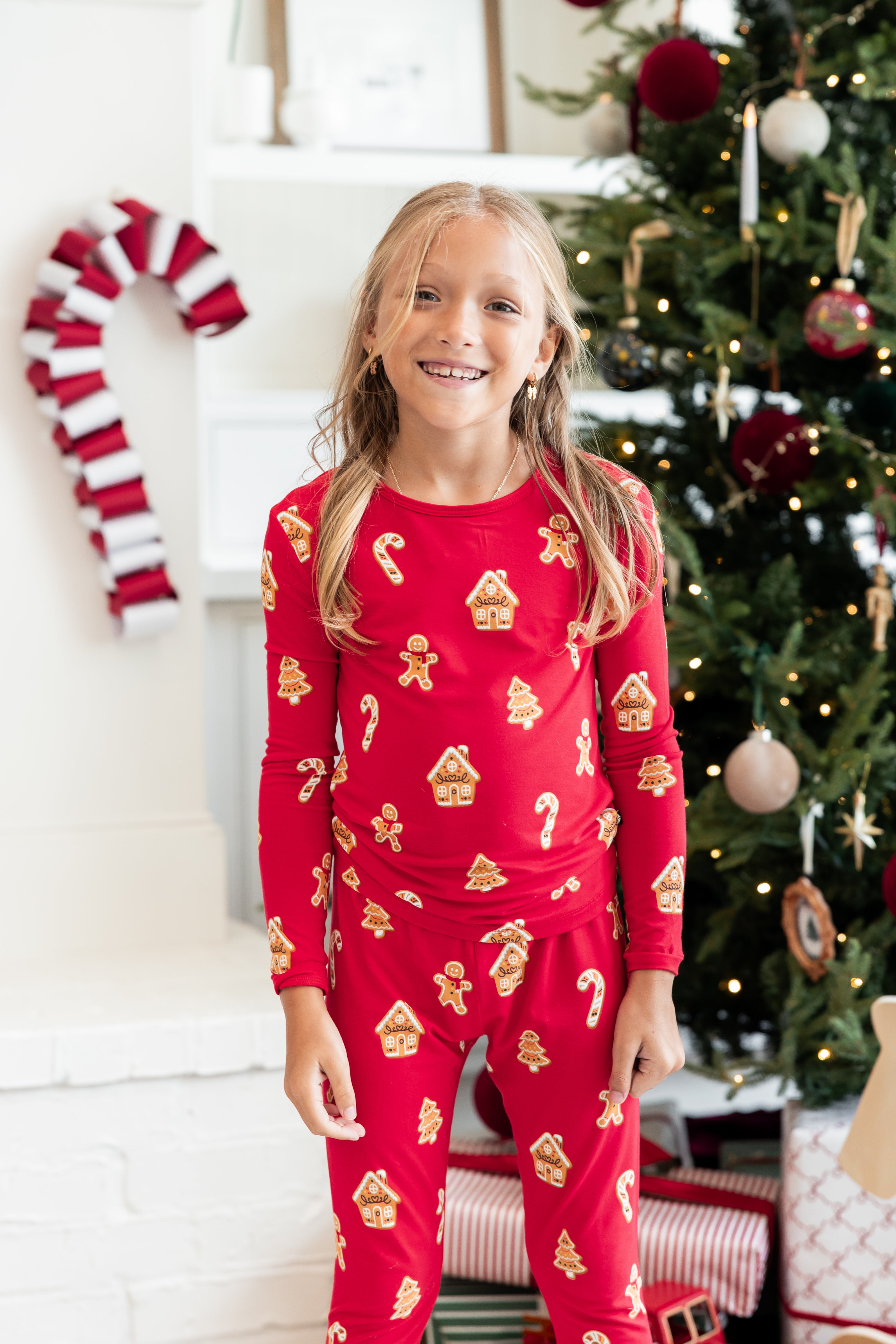 A girl smiling in a festive holiday setting, wearing a red long-sleeve pajama set with a gingerbread house and candy cane pattern. She is standing in front of a decorated Christmas tree and a white brick fireplace with a candy cane hanging on the left.