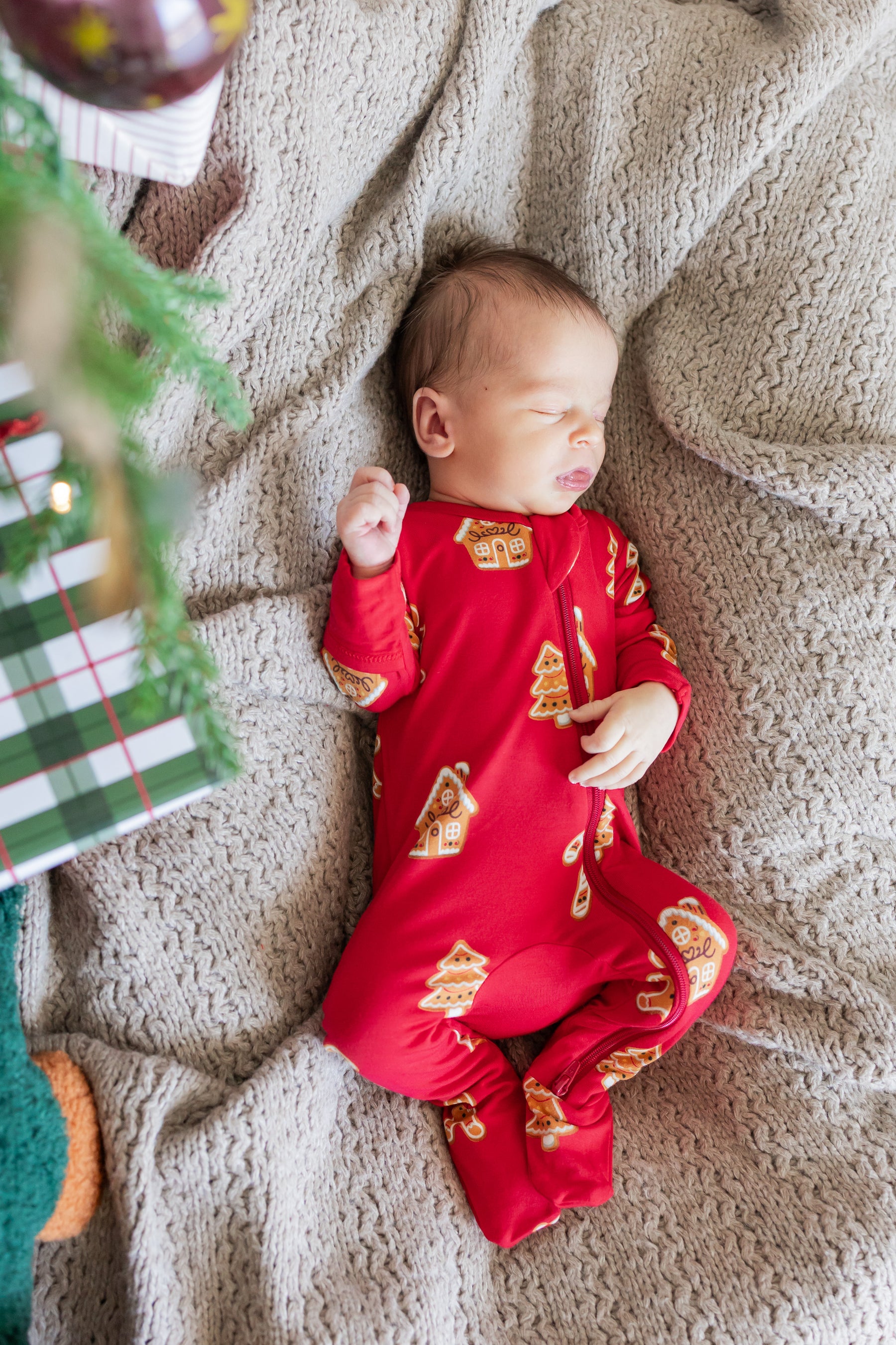 A baby wearing a red footed onesie with a gold Christmas tree pattern, lying on a beige knit blanket next to a wrapped gift.