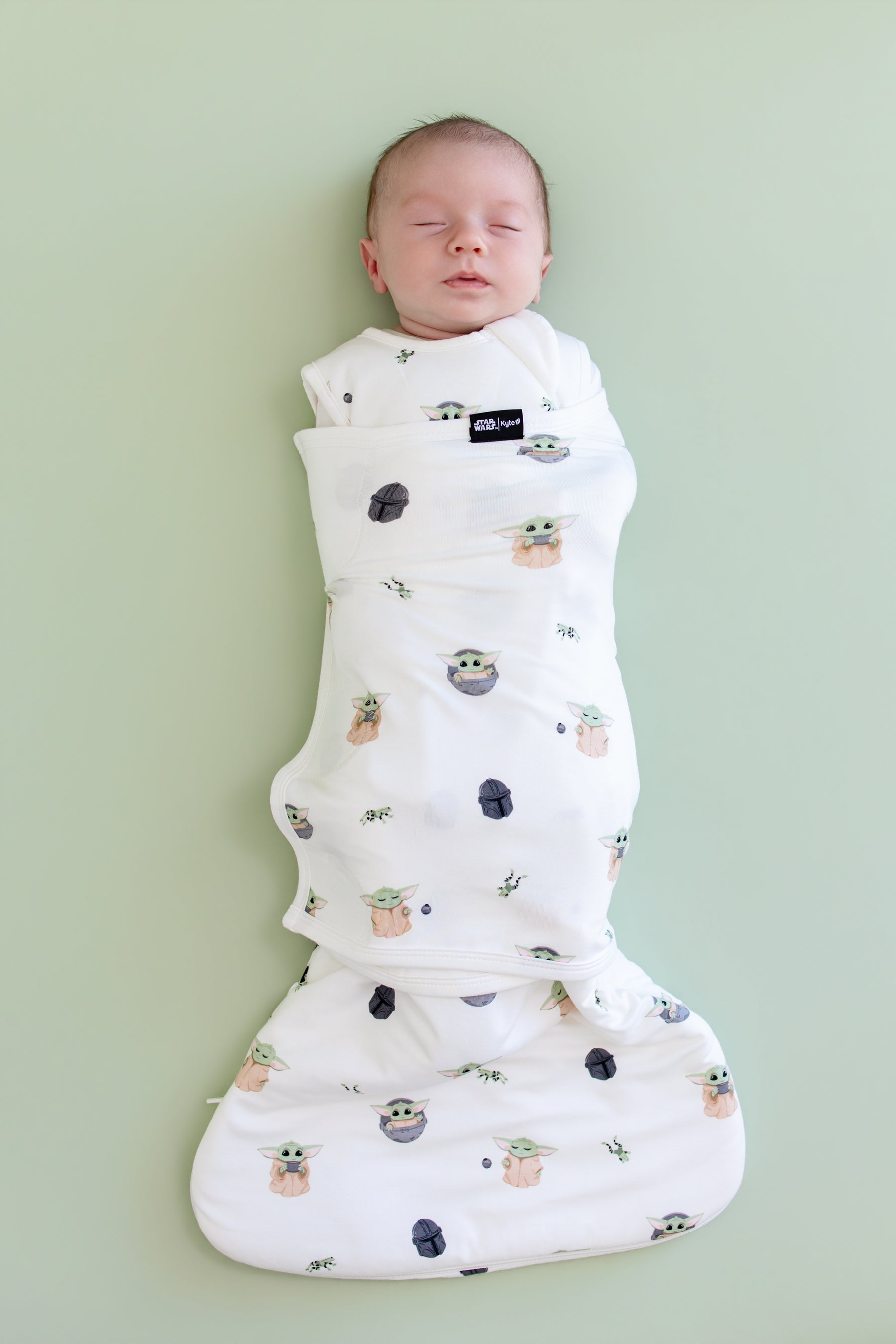 Sleeping newborn swaddled in the Sleep Bag Swaddler in Grogu™ 1.0 on a light green background
