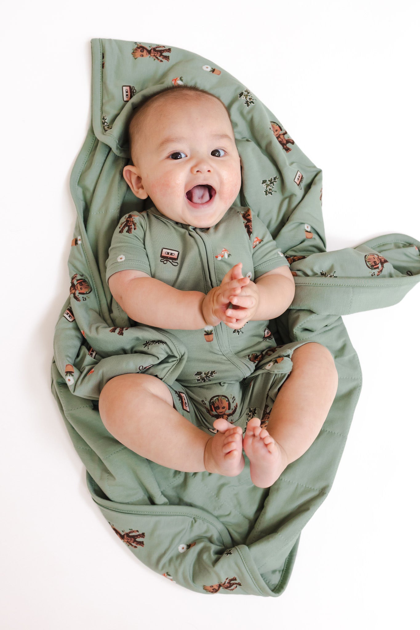 Baby laughing in Green Baby Blanket in Groot 1.0 print wearing a similar print zipper shortall on a white background