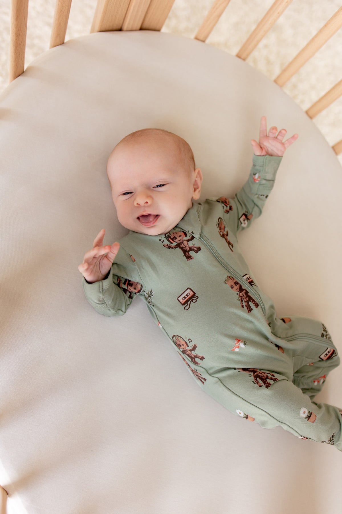 Baby lying in an oval cot wearing Green Zippered Footie in Groot print on a beige bassinet sheet