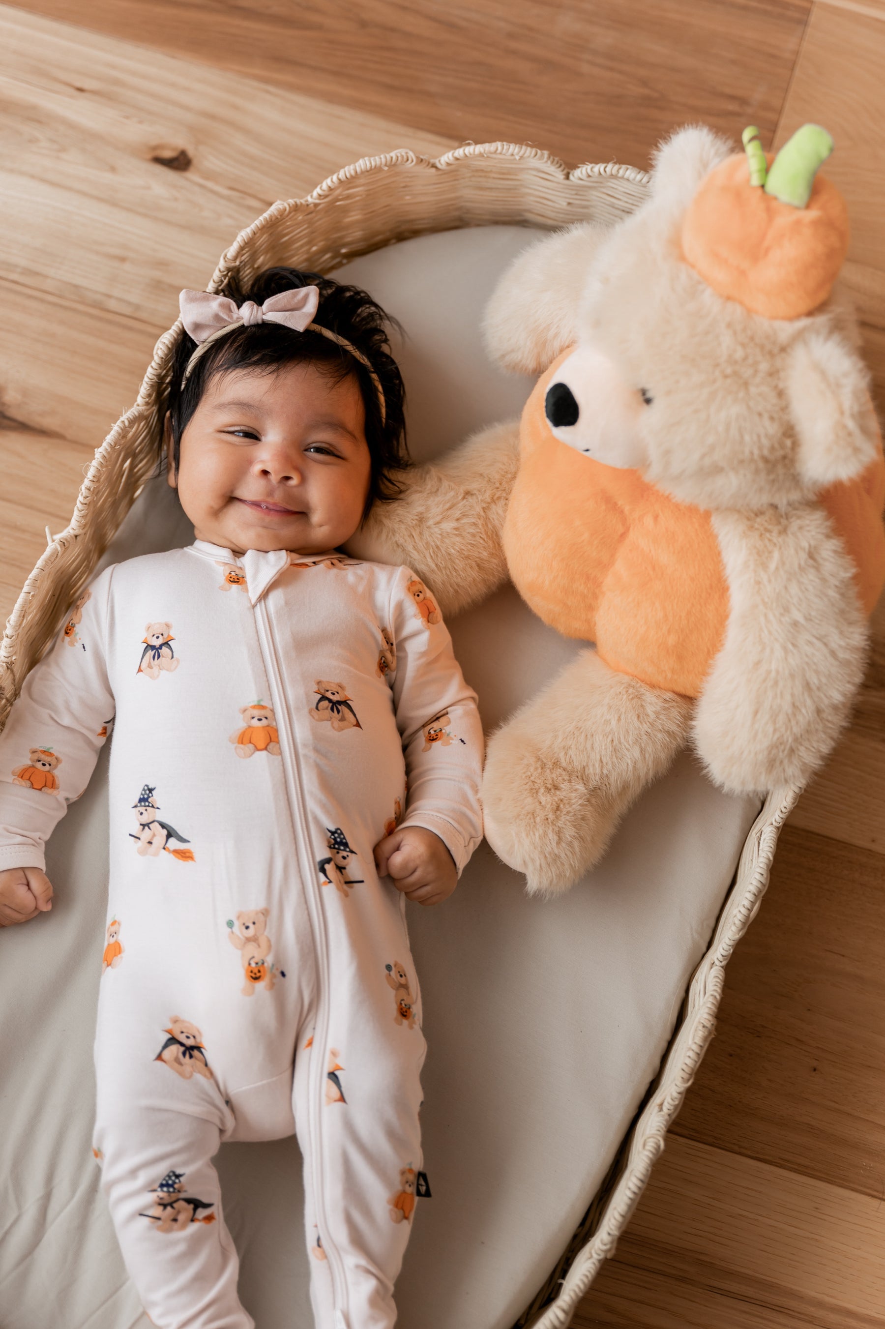 Baby wearing a halloween printed footie with a plush teddy bear dressed as a pumpkin next to her 