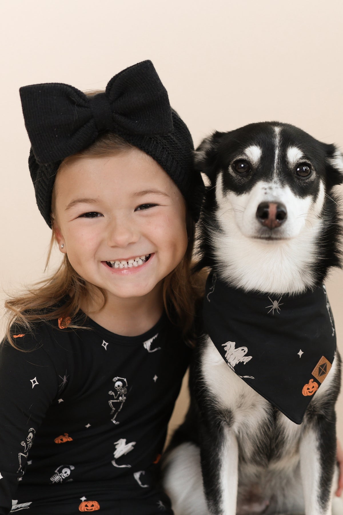 Black and White dog wearing Dog Bandana in Halloween Friends print sitting with a girl in similar print long sleeve pajama set on a beige background