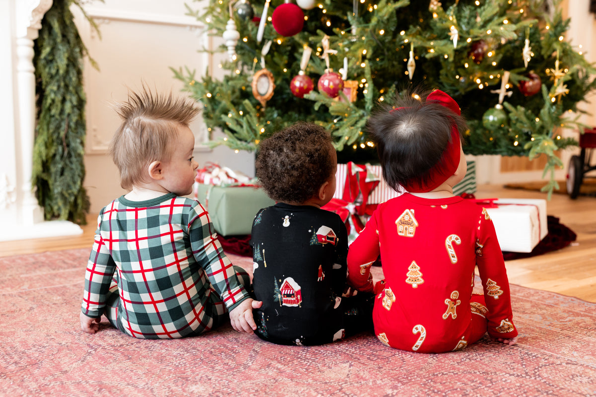 3 babies sitting by a christmas tree wearing festive holiday printed pajamas