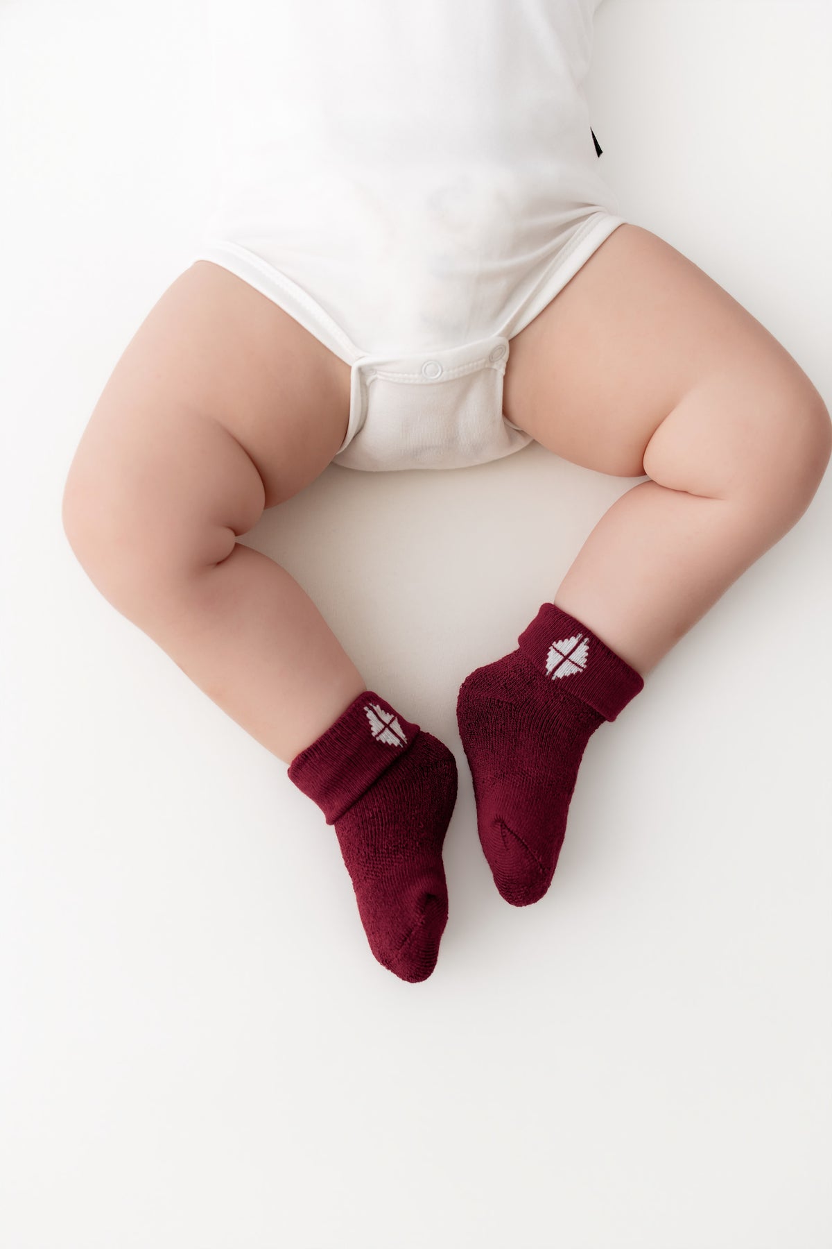 A baby wearing burgundy knit socks with a small white geometric diamond logo on the cuff, shown from a top-down view on a white background.