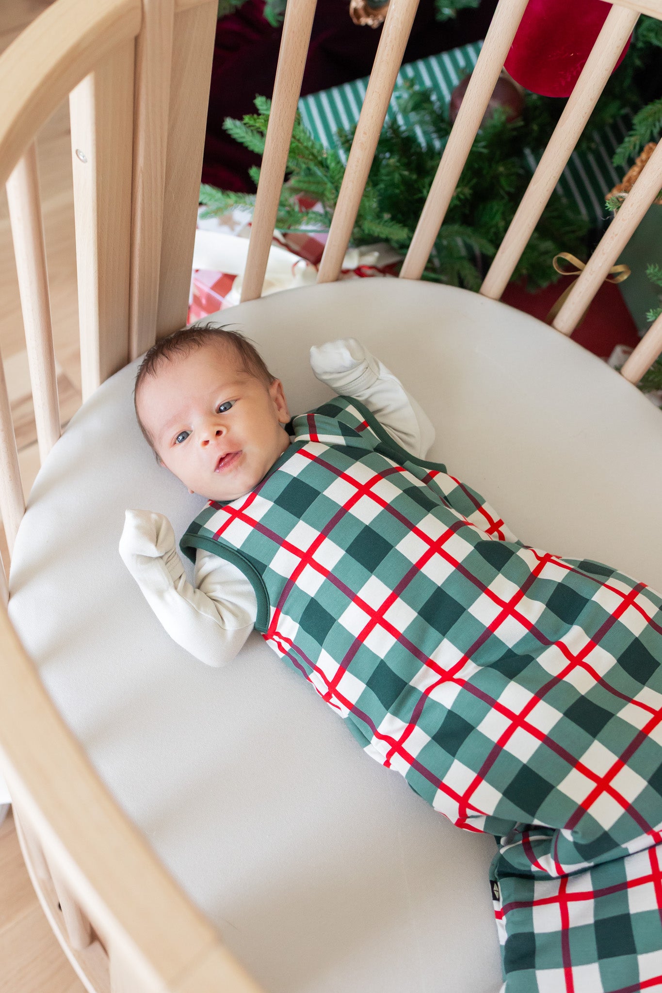 A baby lying on their back in a light wood crib, wearing a green, white, and red plaid sleep sack. A Christmas tree is visible in the background.