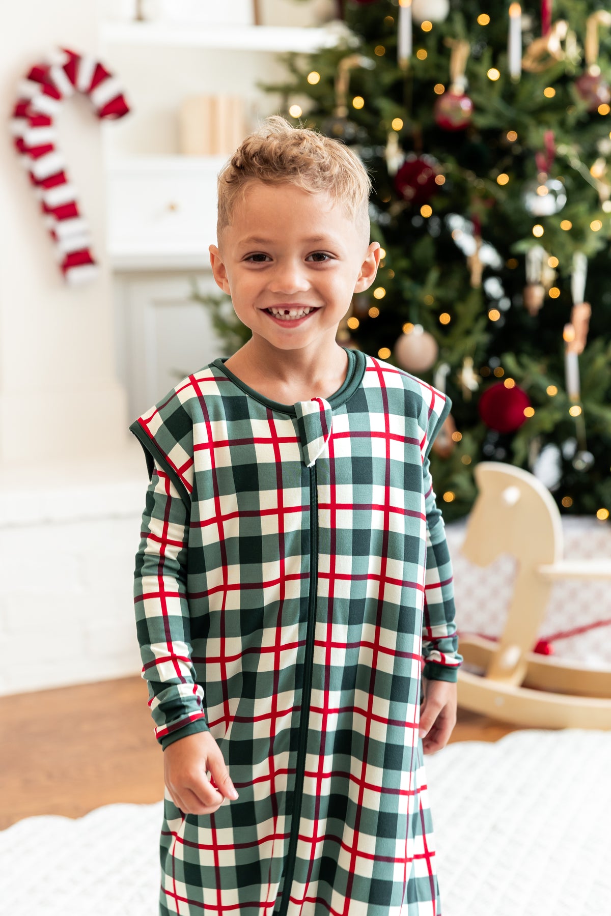 A young boy smiling in a festive holiday room, wearing a green, white, and red plaid zip-up sleep sack walker. A decorated Christmas tree, a wooden rocking horse, and a candy cane are visible in the softly blurred background.