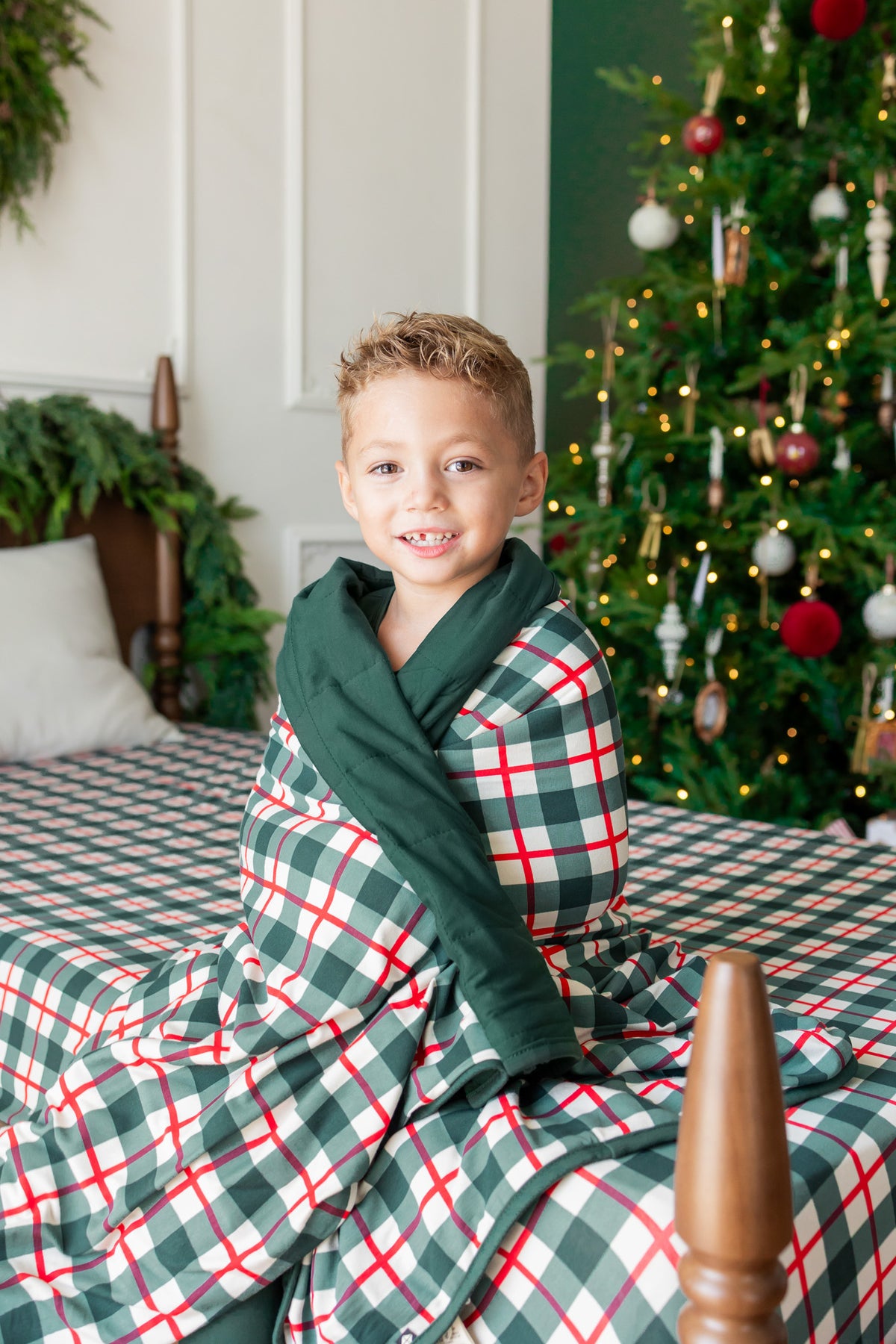 A young boy smiling while wrapped in a green Christmas-themed plaid blanket on a bed with a decorated Christmas tree in the background.