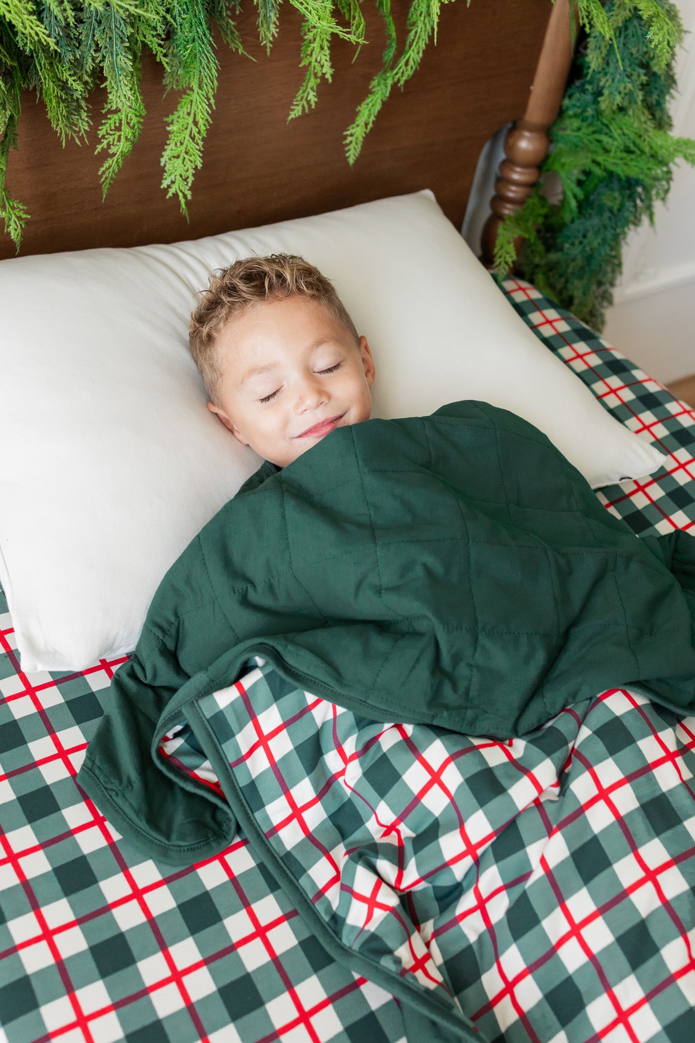 A child sleeping soundly on a bed, covered with a dark green blanket, under a green and red plaid holiday-themed sheet. The background features a wooden headboard decorated with green garlands.