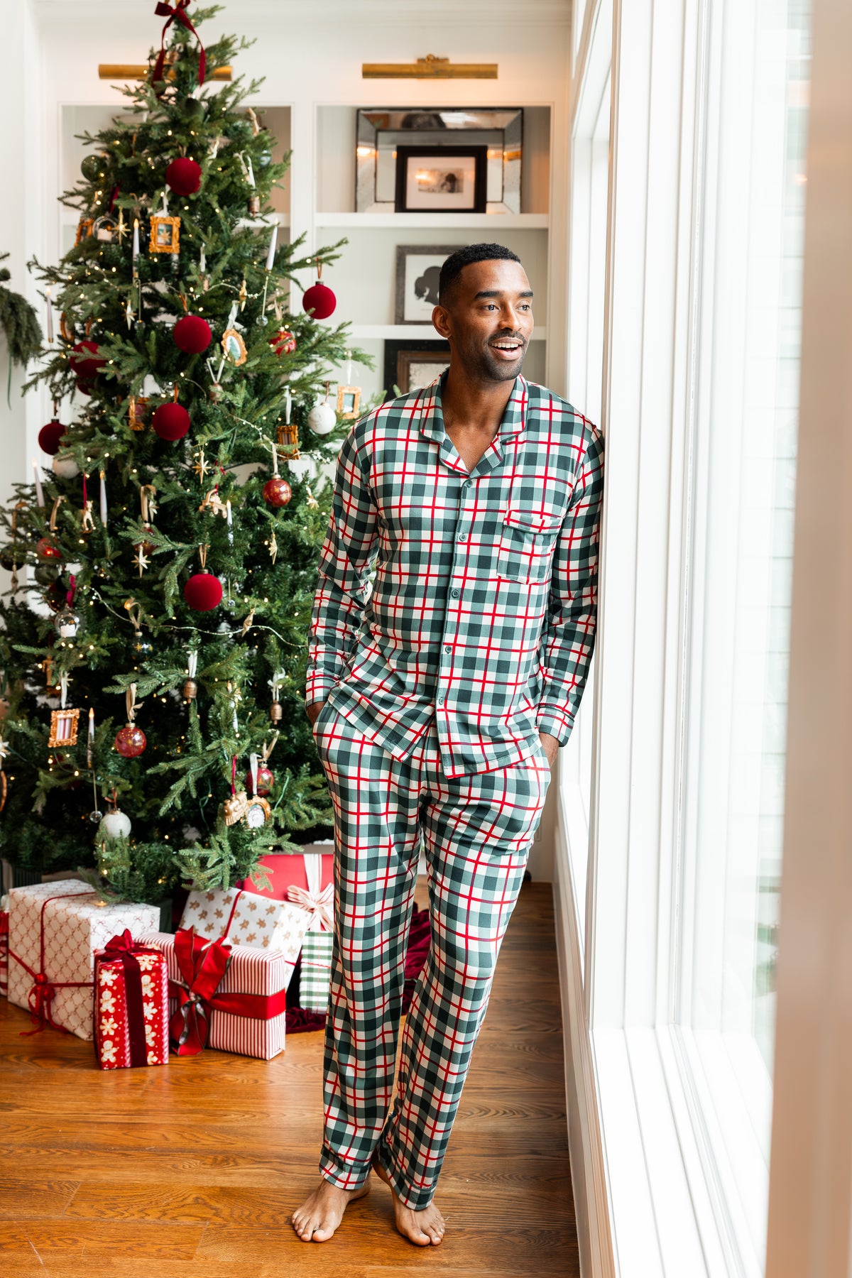 A man wearing a green, red, and white plaid pajama set stands barefoot in a home hallway next to a large window, with a decorated Christmas tree and wrapped gifts in the background.